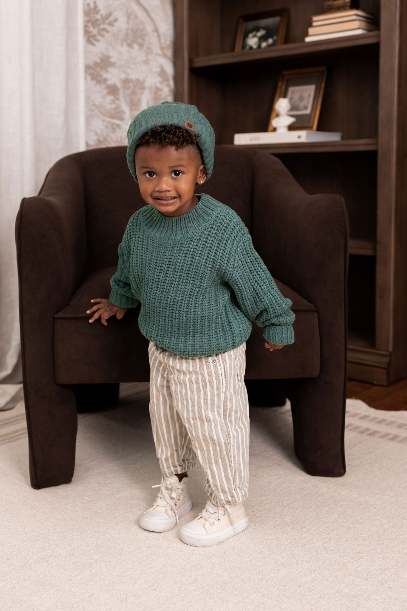 Boy wearing Chunky Knit Sweater and chunky knit ribbed beanie in Pine over beige pants standing in a living room