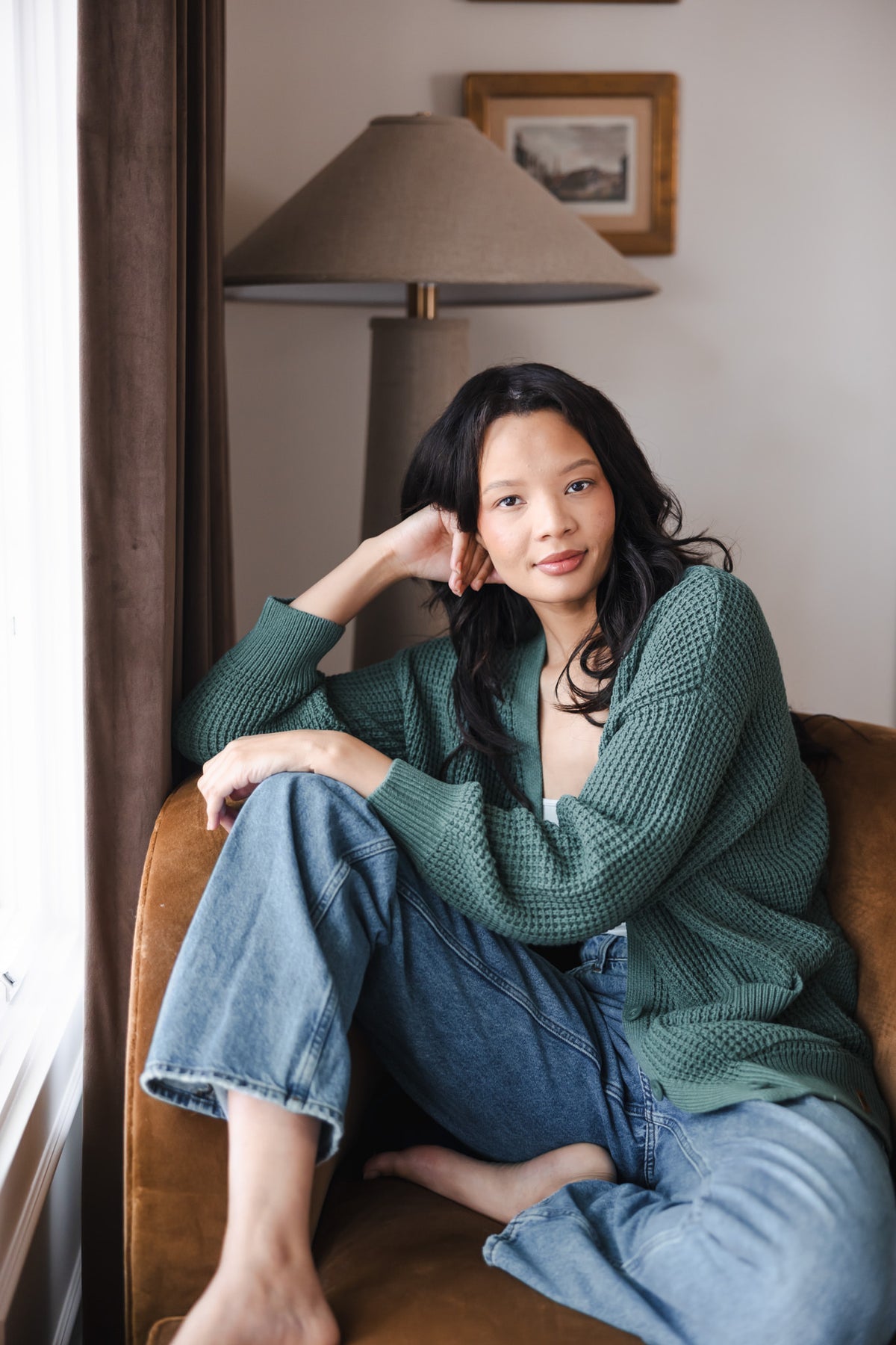 Modal sitting on a brown sofa posing in Chunky Knit Women's Waffle Cardigan in Pine wearing it over blue jeans