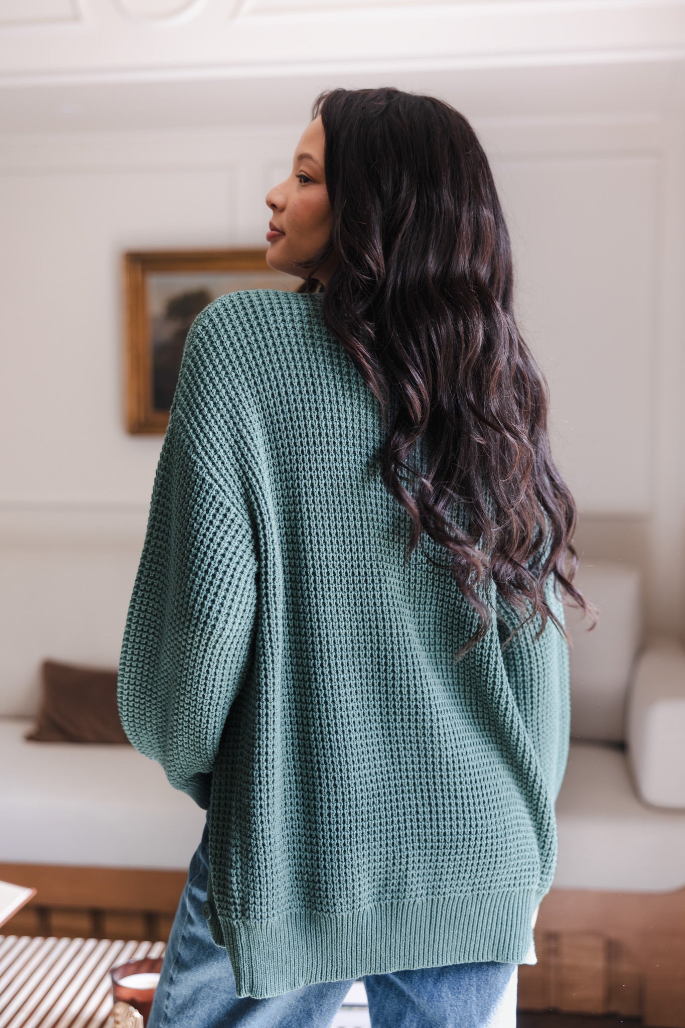 Modal showcasing the back side of Chunky Knit Women's Waffle Cardigan in Pine over blue jeans standing in a living room