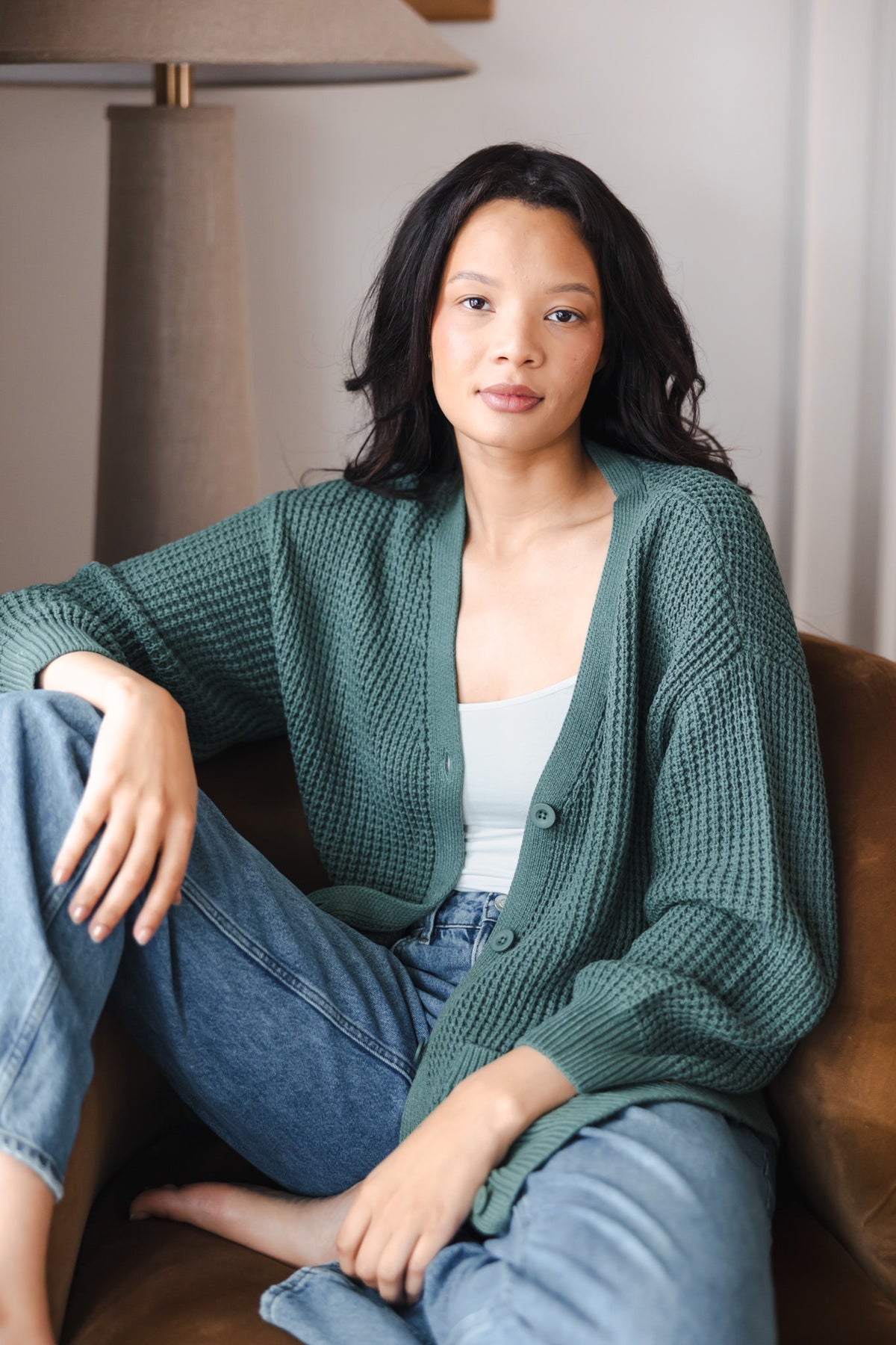 Modal posing in Chunky Knit Women's Waffle Cardigan in Pine over white camisole and blue jeans sitting on a brown sofa