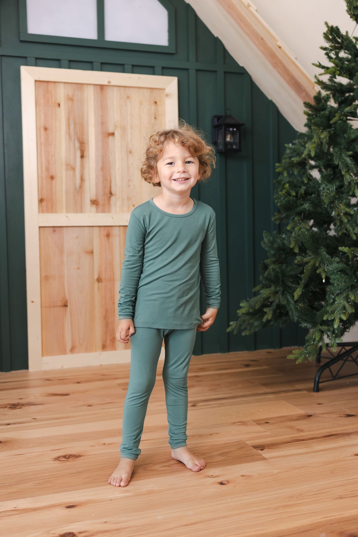 A young child wearing a matching sage long sleeve pajama set. The background features dark green paneled walls, a light wood door, and a Christmas tree on the right.