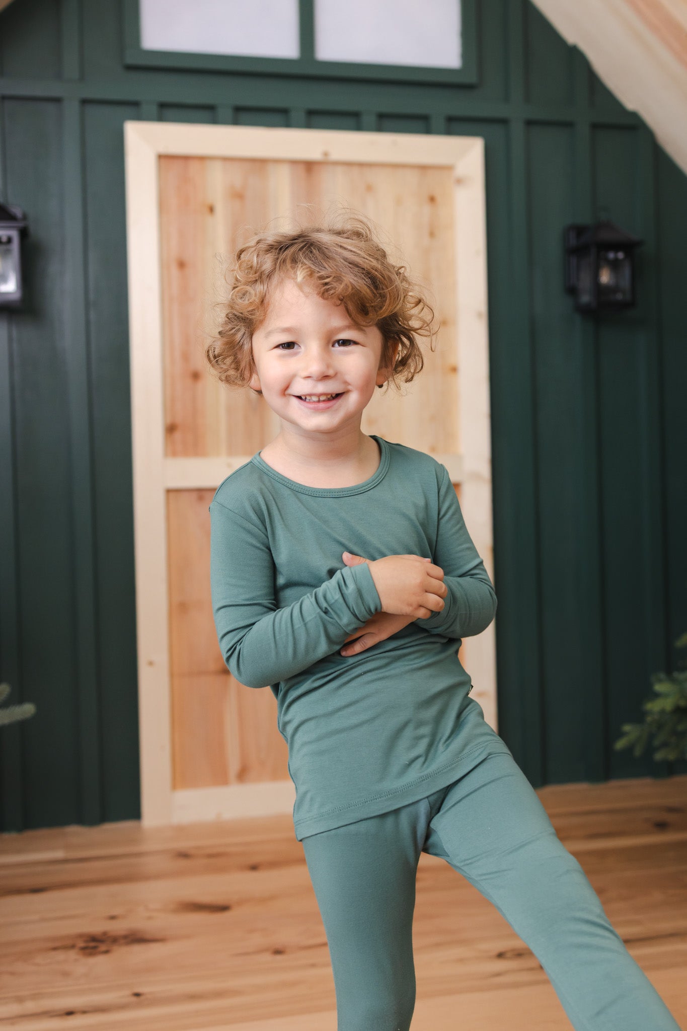 A young child wearing a matching sage green long-sleeve pajama set, standing on a wooden deck in front of a dark green house wall with a light wood door.