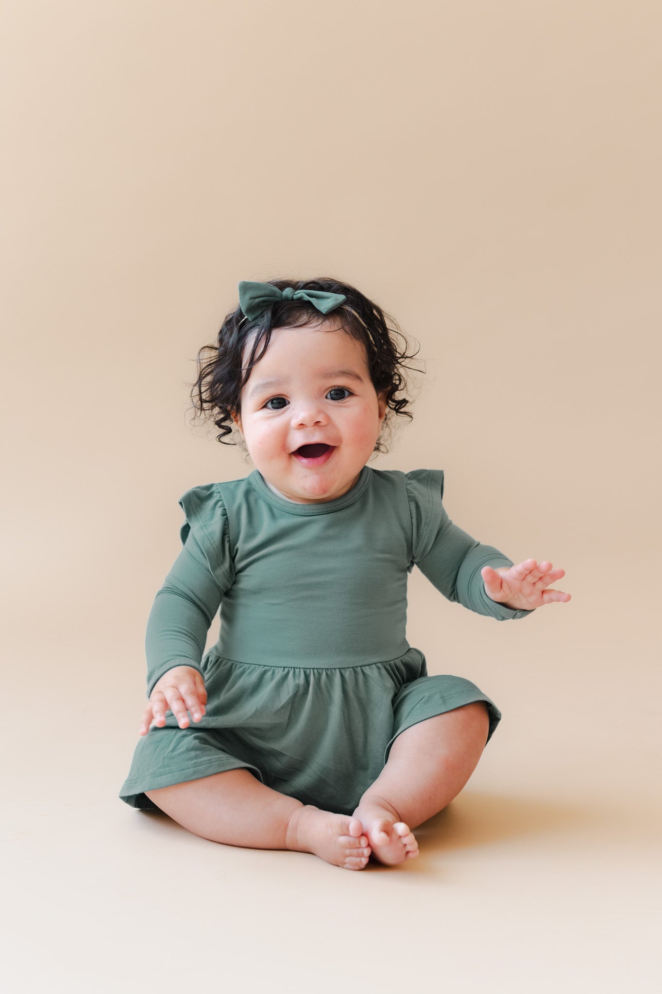A smiling baby girl sitting on a warm beige studio background, wearing a sage green long-sleeve bodysuit dress with ruffled shoulders and a matching green bow headband.