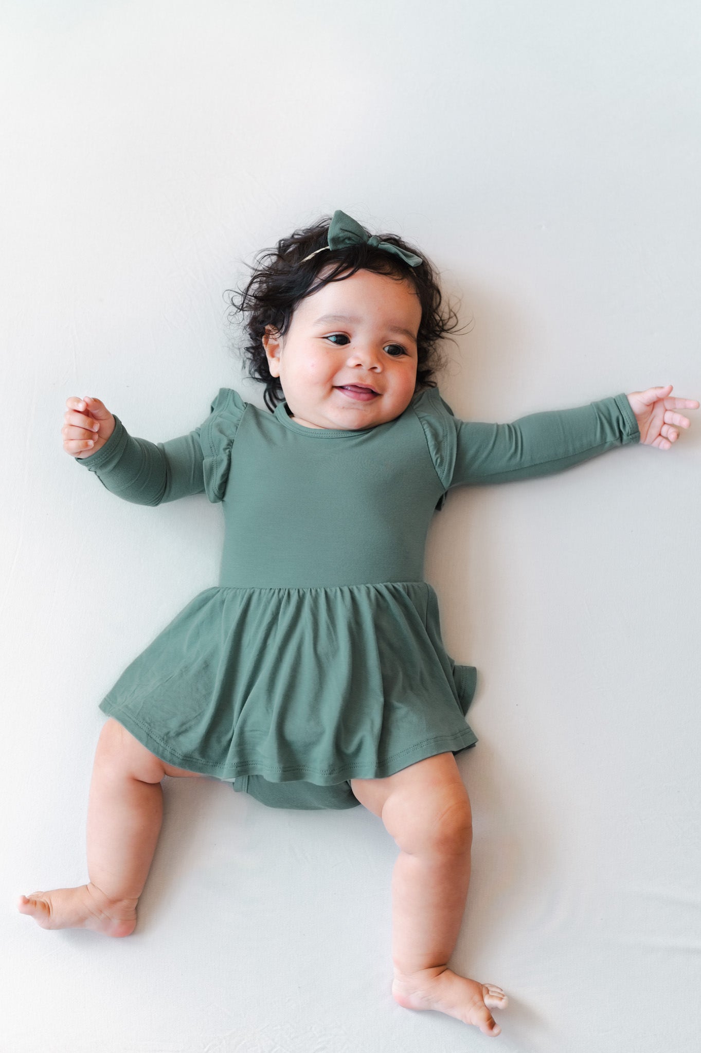 A baby lying on a light gray background wearing a sage green long-sleeve dress with a ruffled skirt and a matching green bow headband.
