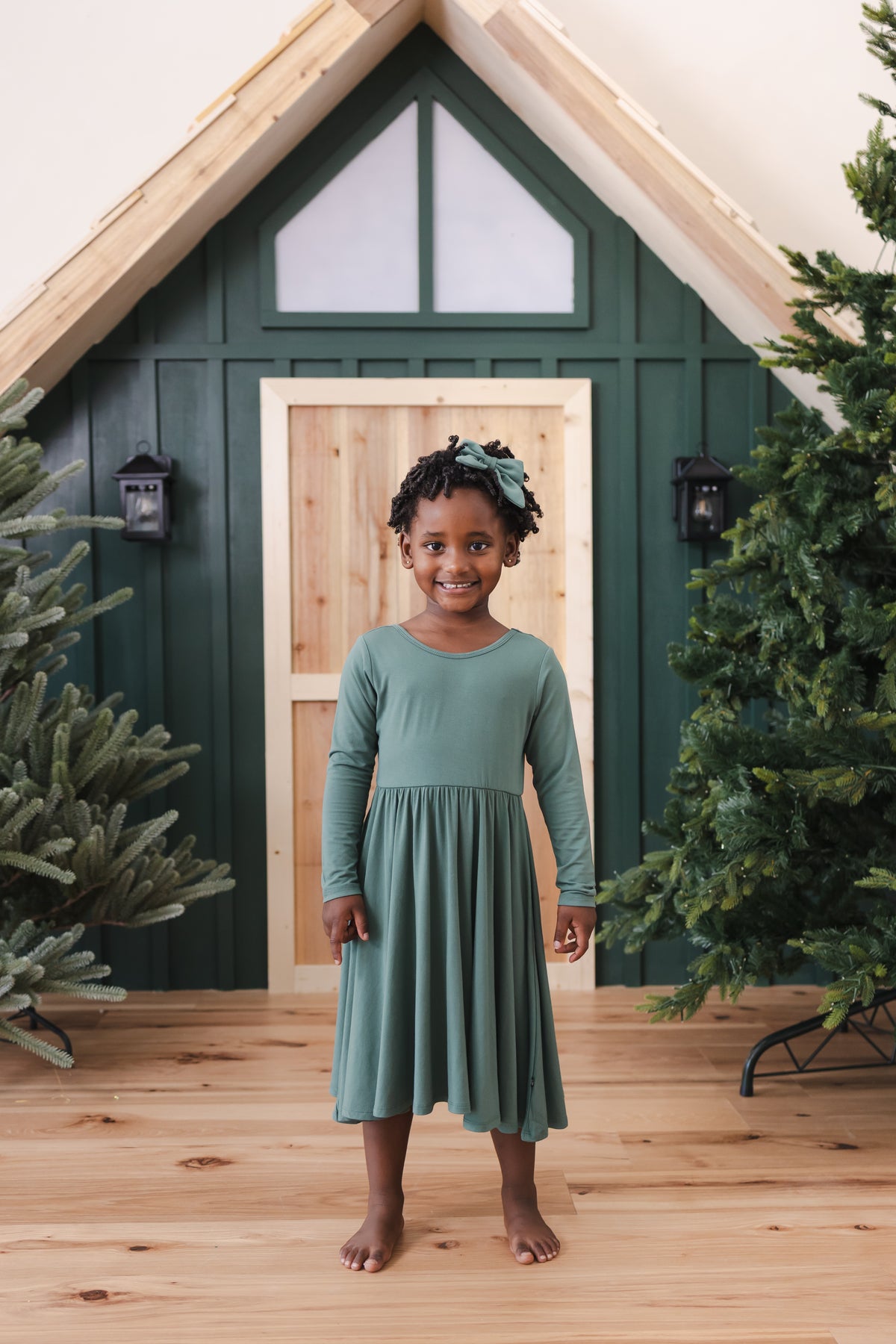 Girl smiling in Long Sleeve Twirl Dress in Pine standing in front of a beige door