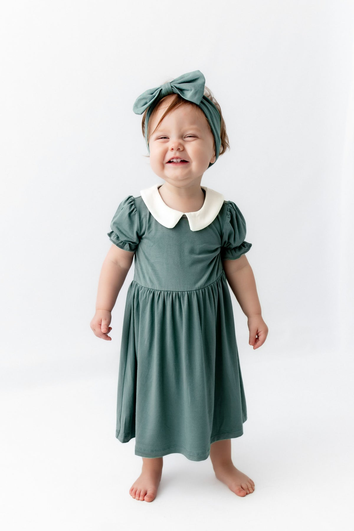 A toddler girl wearing a sage green dress with a white Peter Pan collar, matching green bow headband, and standing barefoot against a white studio background.