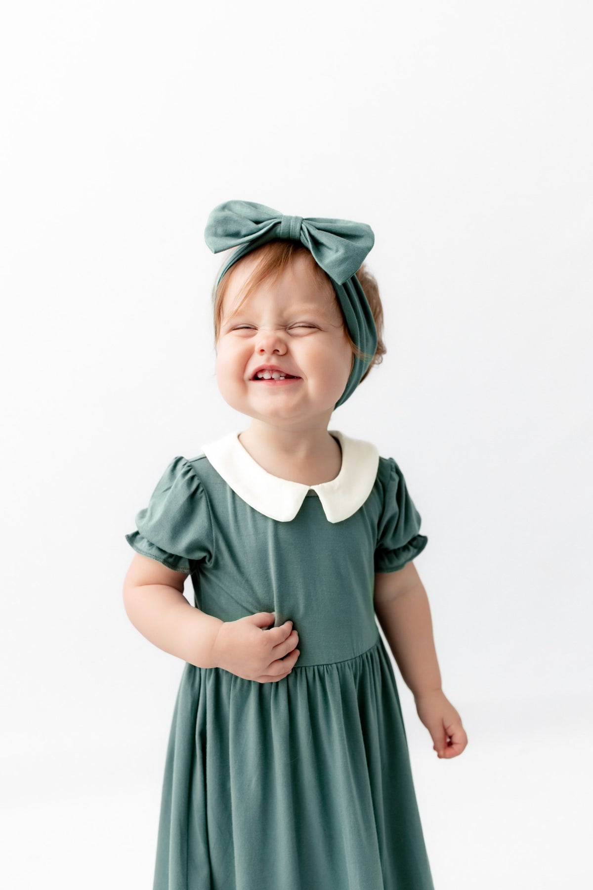 A toddler wearing a sage green dress with a white Peter Pan collar and a matching sage green bow headband, smiling against a plain white background.