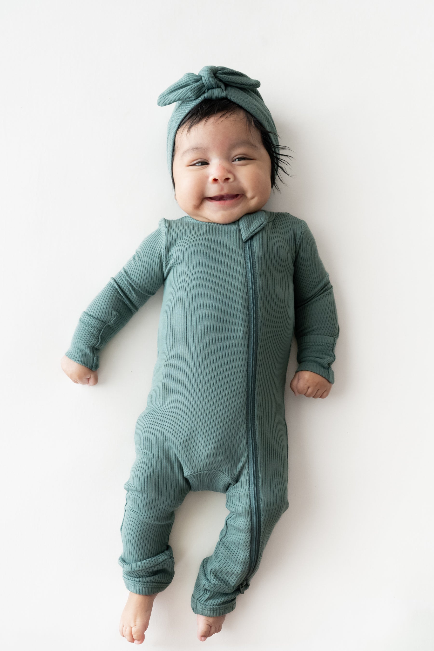 Baby wearing Ribbed Headwrap in Pine with zippered romper on a beige background
