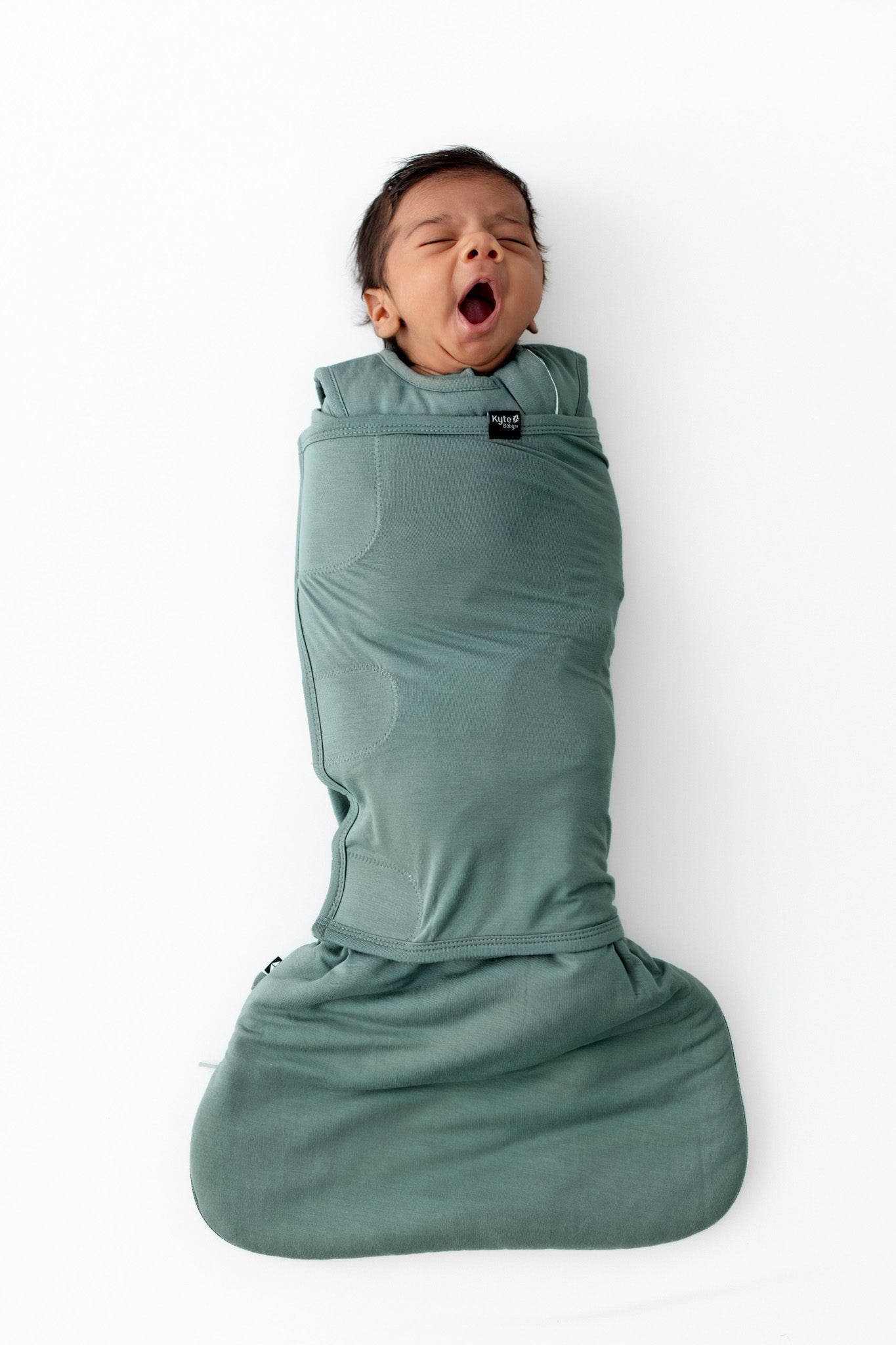 A baby wrapped in a sage green swaddler, yawning while lying on a white background.