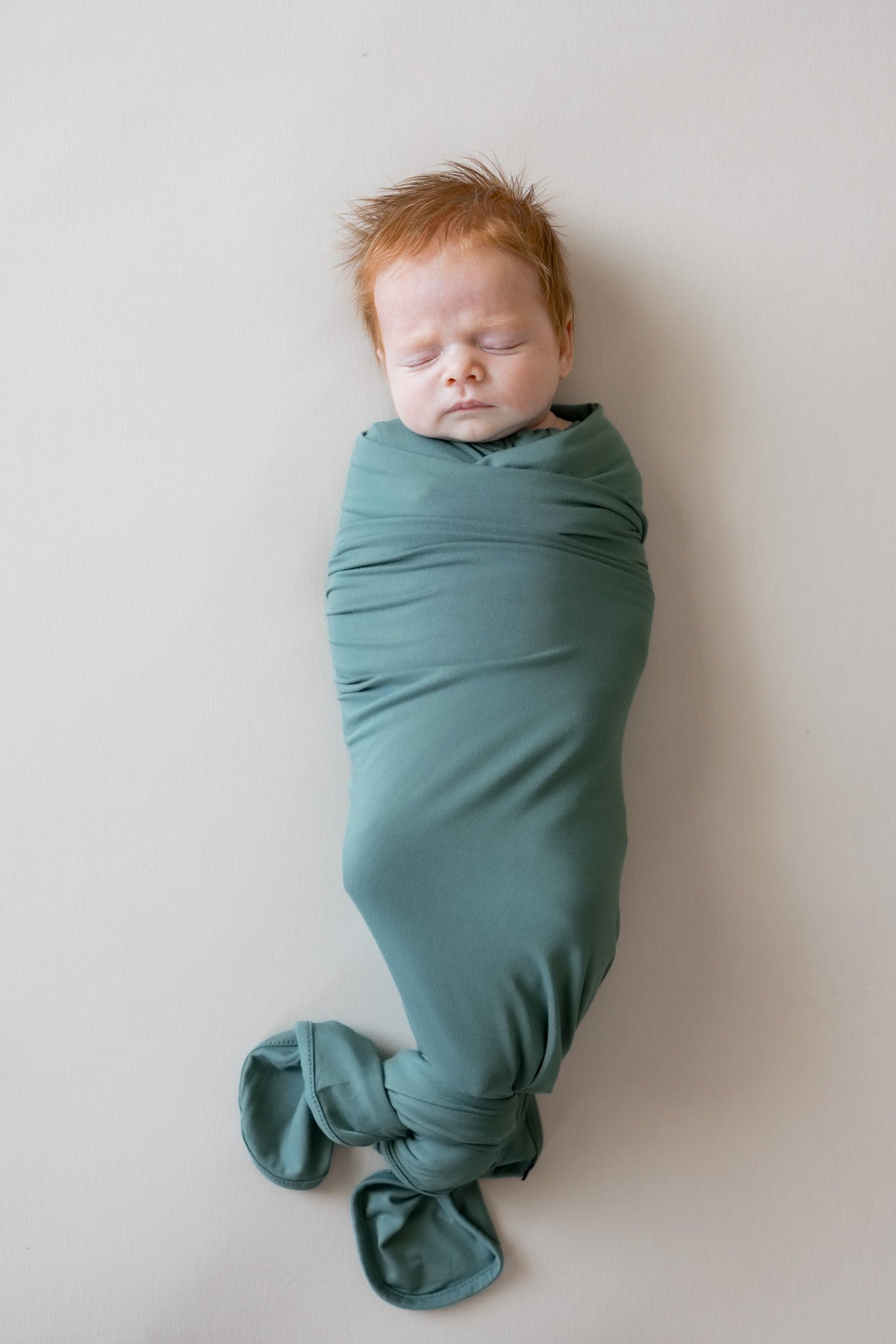 A sleeping newborn baby swaddled in a dusty green fabric wrap, lying on a neutral light beige background.