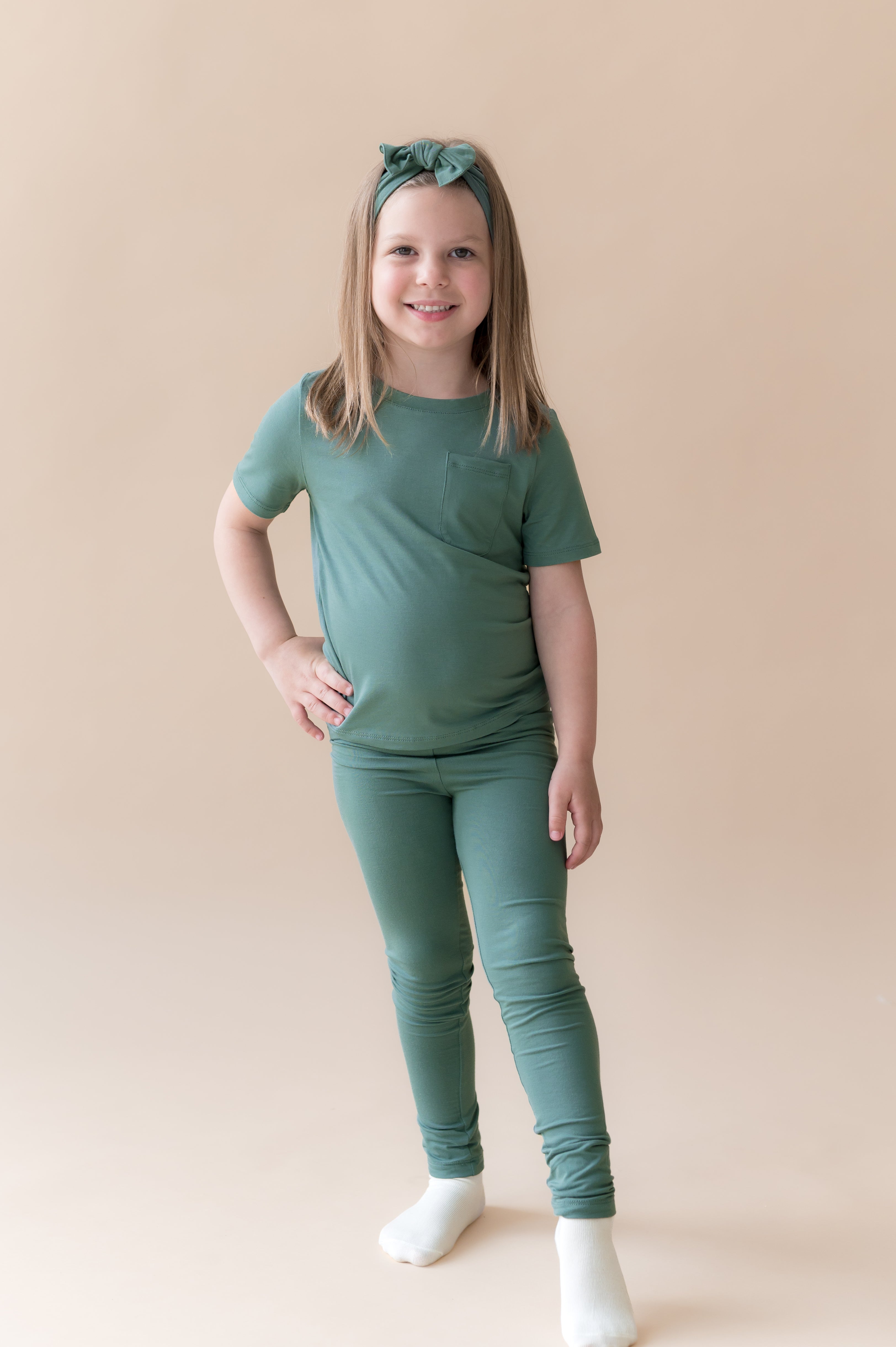 Model wearing Toddler Leggings in Pine under pine shirt with knotted bow headband in pine on a beige background