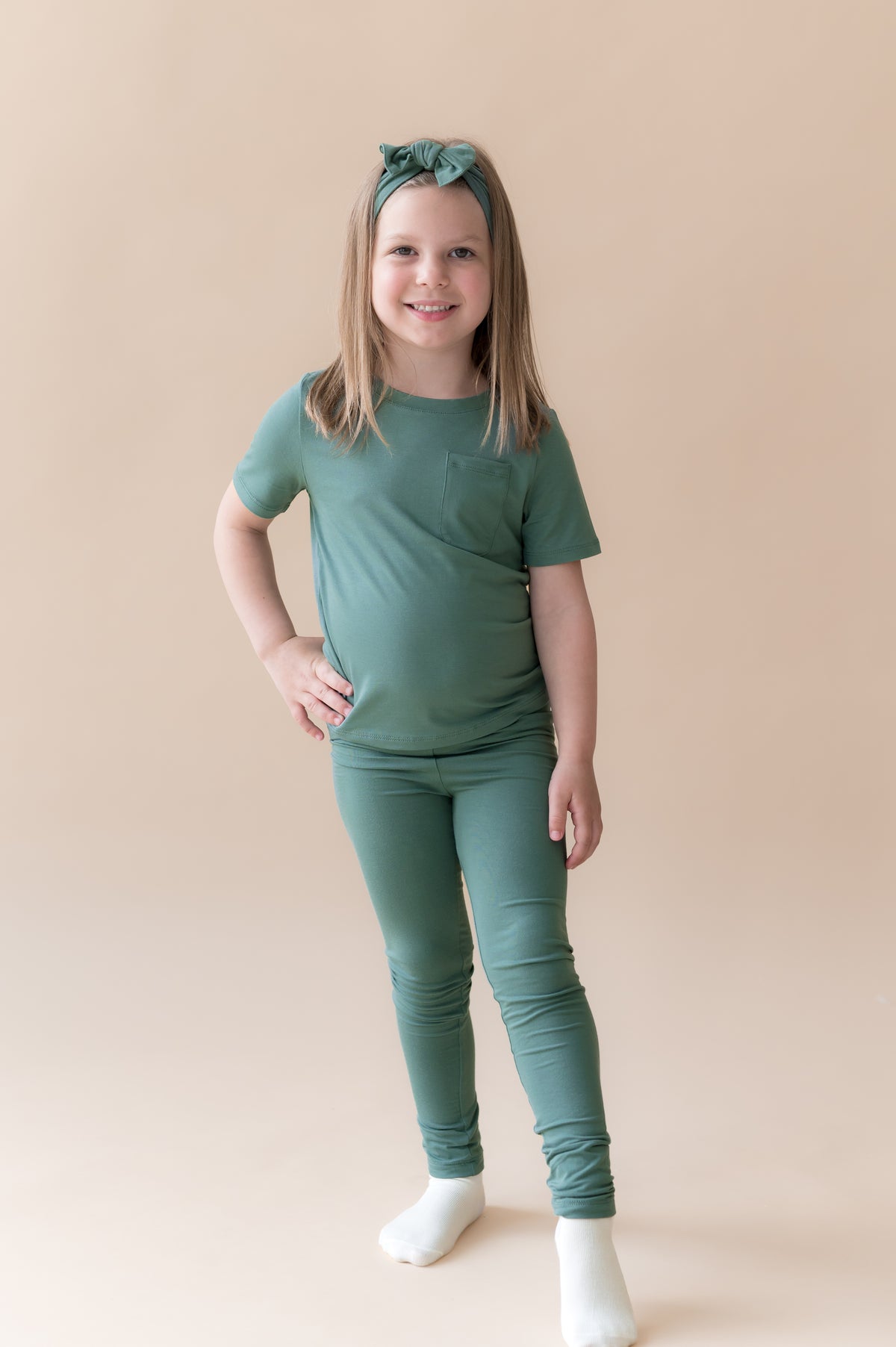 Model wearing Toddler Leggings in Pine under pine shirt with knotted bow headband in pine on a beige background