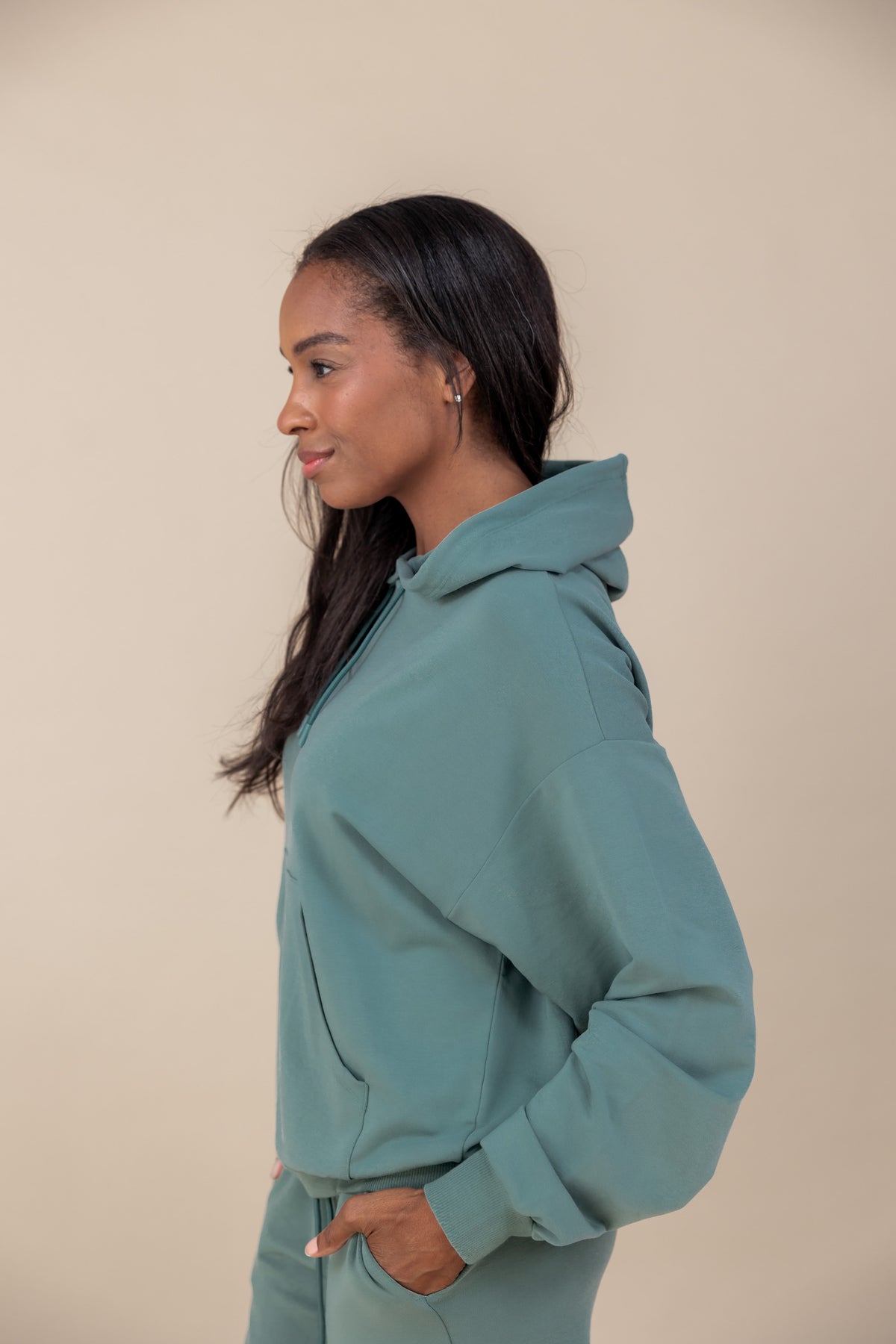 Side pose of woman wearing Women's Hoodie in Pine color over a similar color jogger pants on a beige background