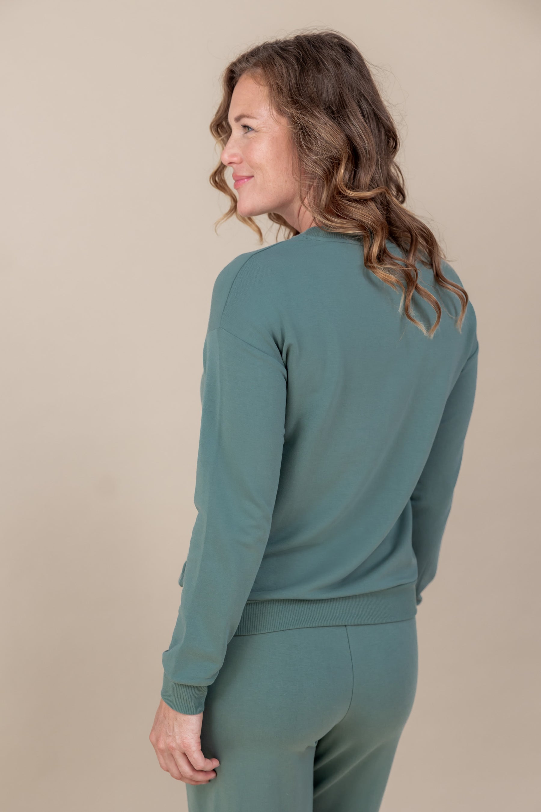 Back side of Woman wearing Women's Sweatshirt in Pine color over Women's sweatpants in pine on a beige background
