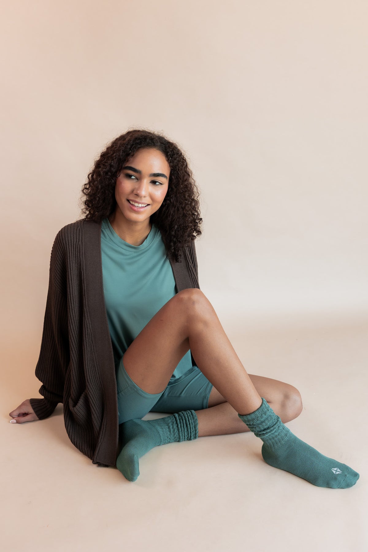Model sitting on a beige background wearing Women’s Biker Short Set in Pine and Adult Socks in Pine with Chunky Knit Oversized Cardigan in Espresso