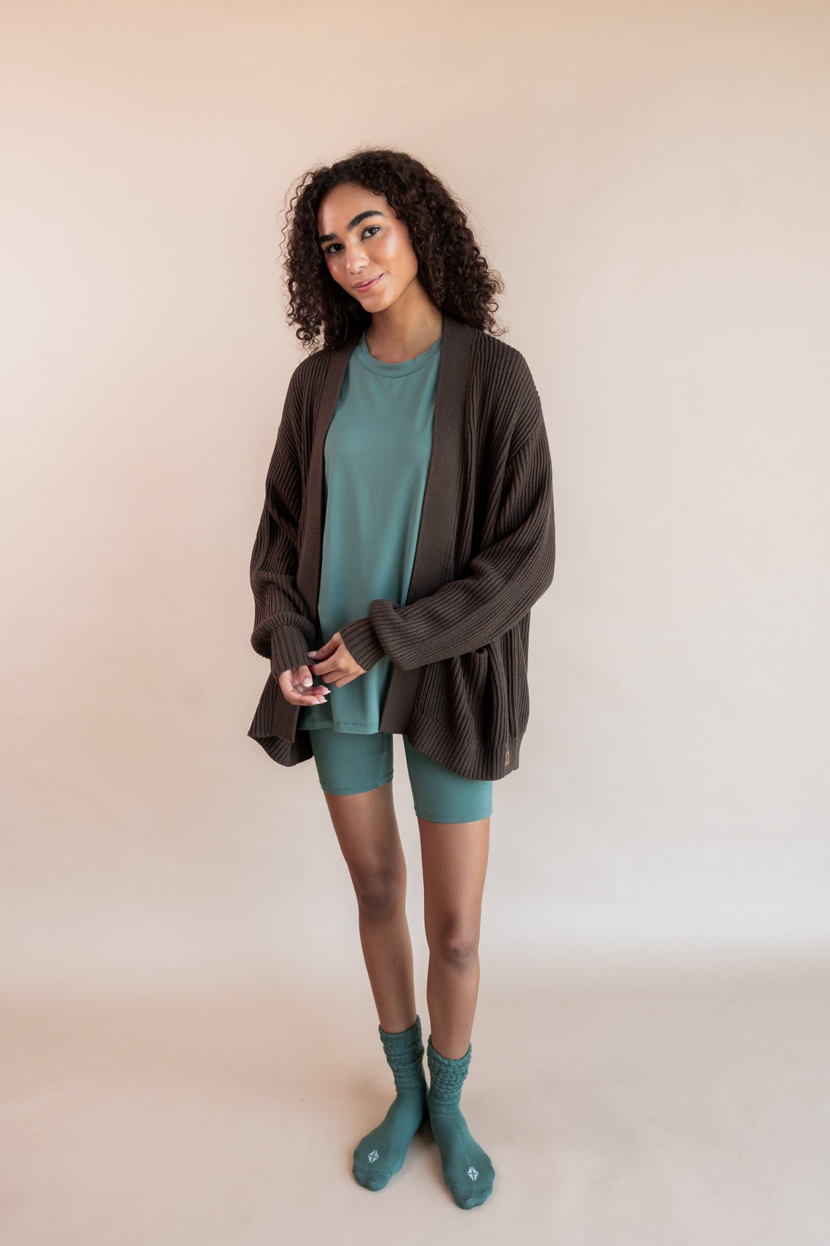 Model wearing Women’s Biker Short Set in Pine and Adult Socks in Pine with Chunky Knit Oversized Cardigan in Espresso on a beige background