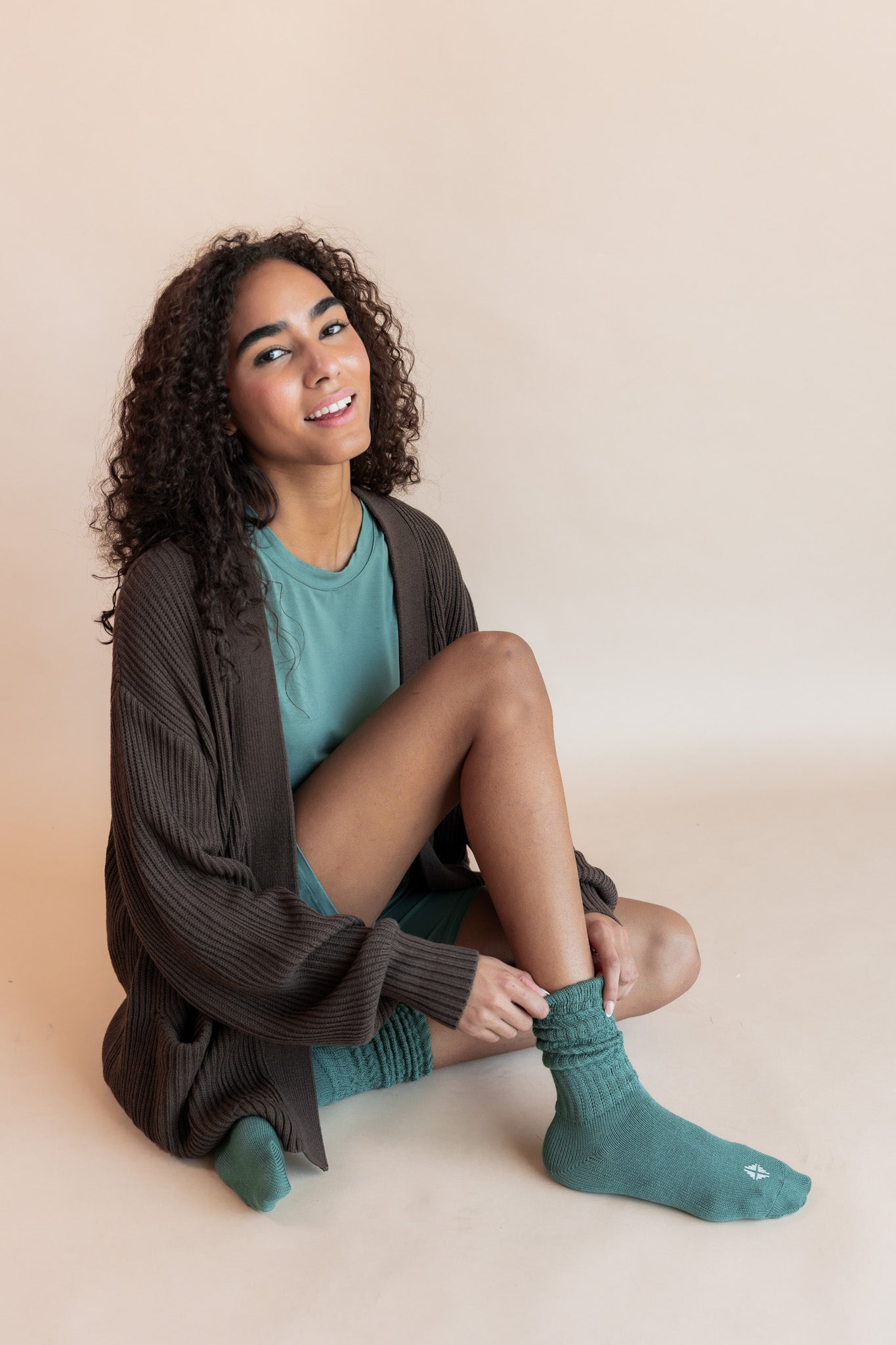 Model sitting on a beige background wearing Women’s Biker Short Set in Pine and Adult Socks in Pine with Chunky Knit Oversized Cardigan in Espresso