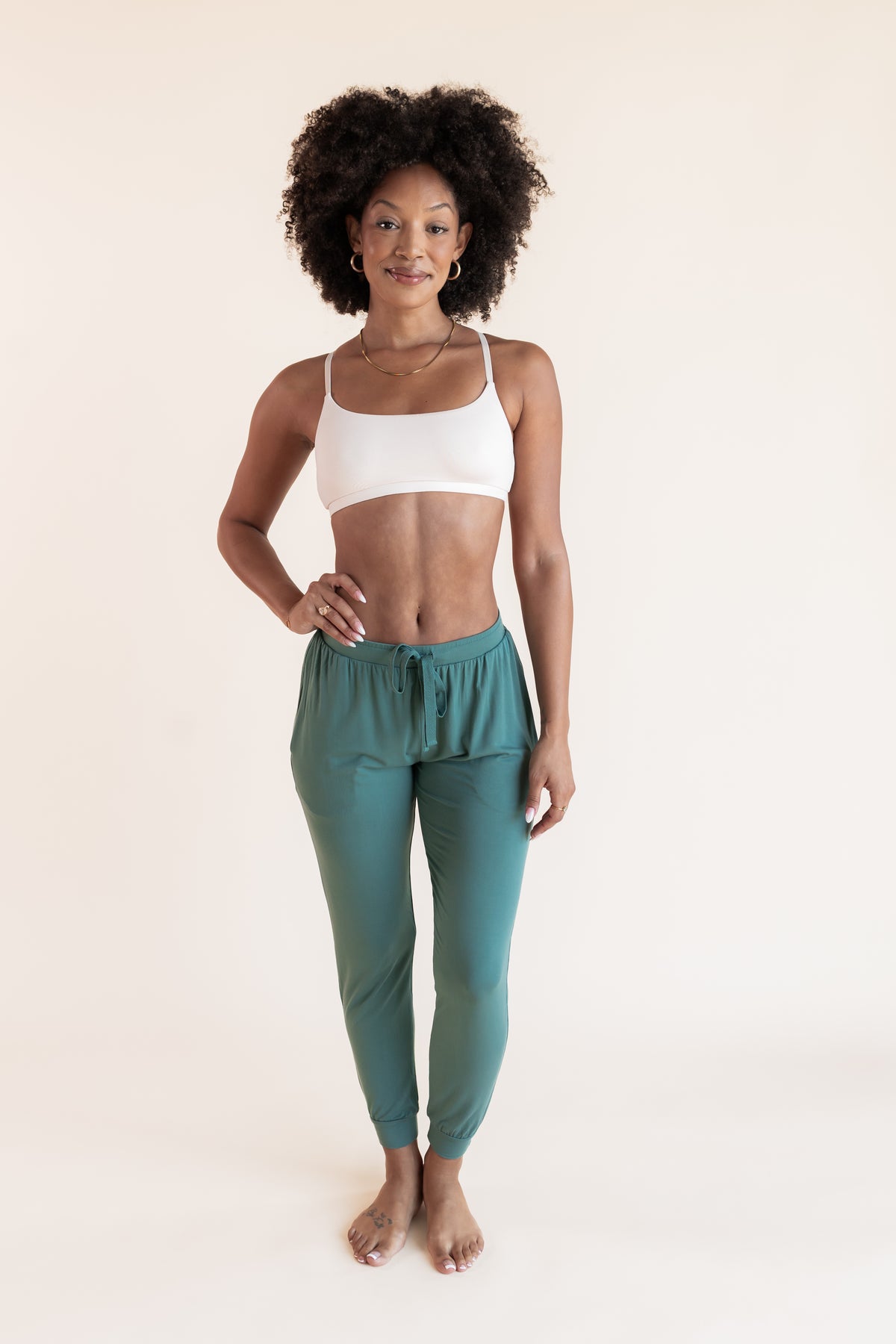 Model posing in Women's Jogger Pants in Pine under white bralette on a beige background