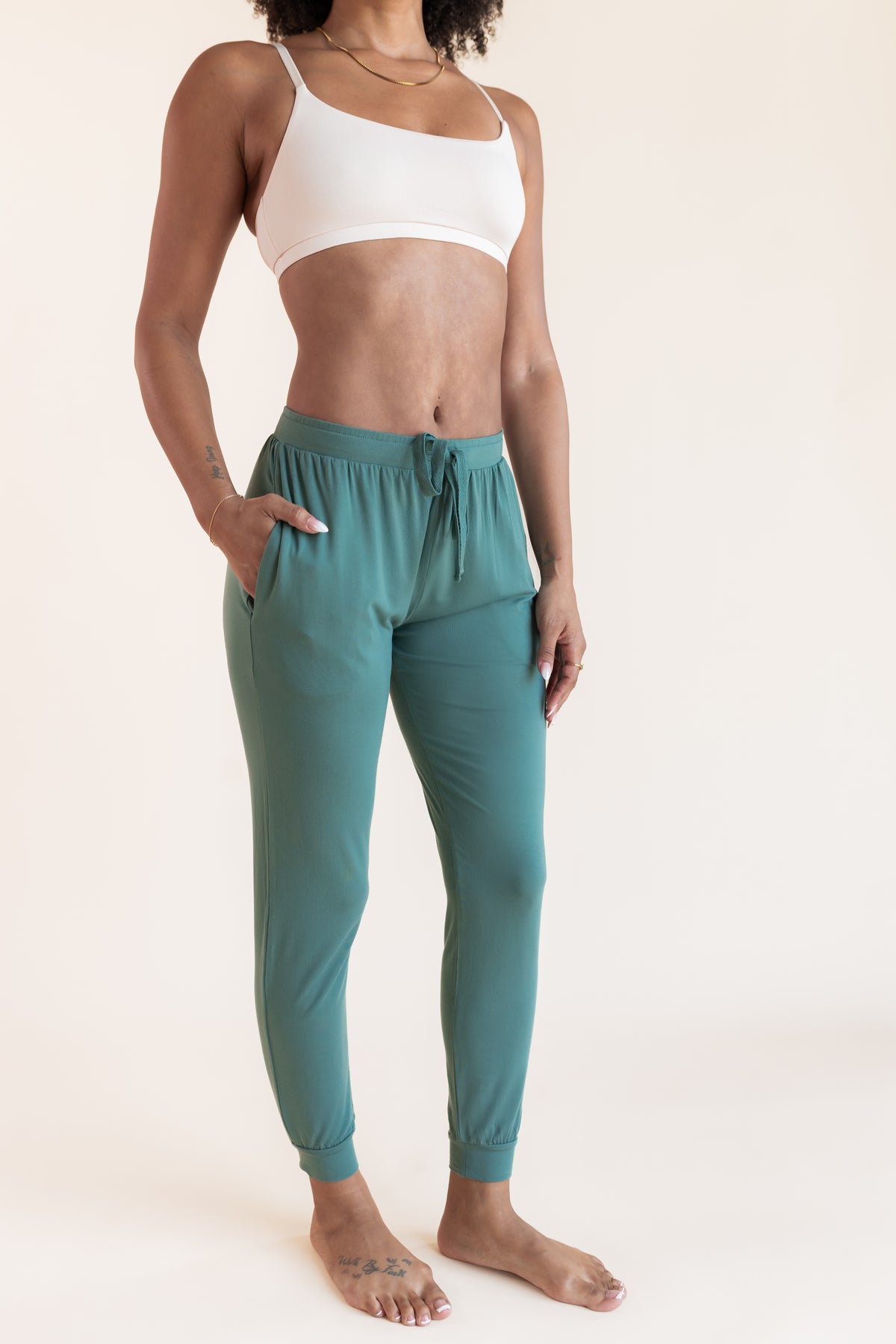 Model wearing Women's Jogger Pants in Pine with pockets and drawstring under white bralette on a beige background