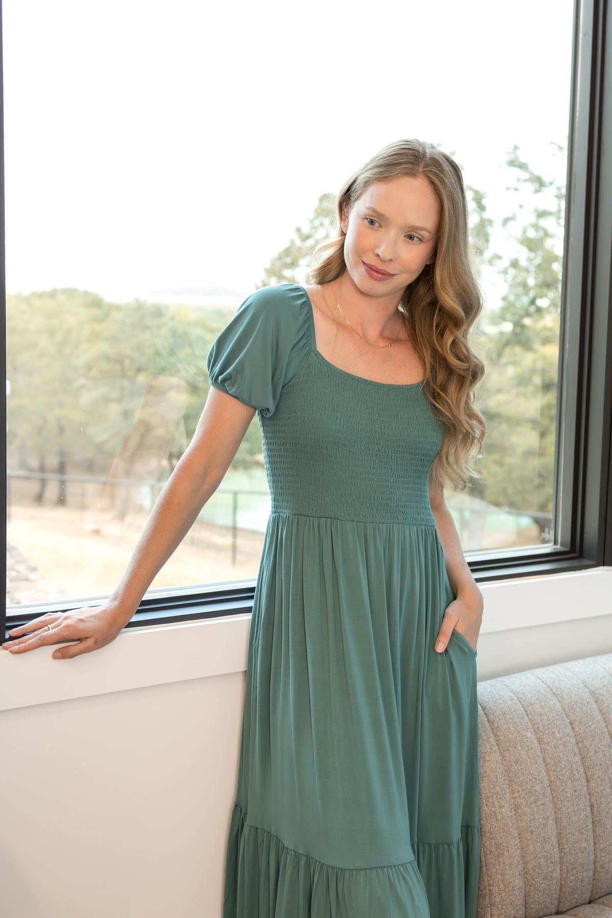 Model posing in Women’s Smocked Dress in Pine with pockets standing in a living room