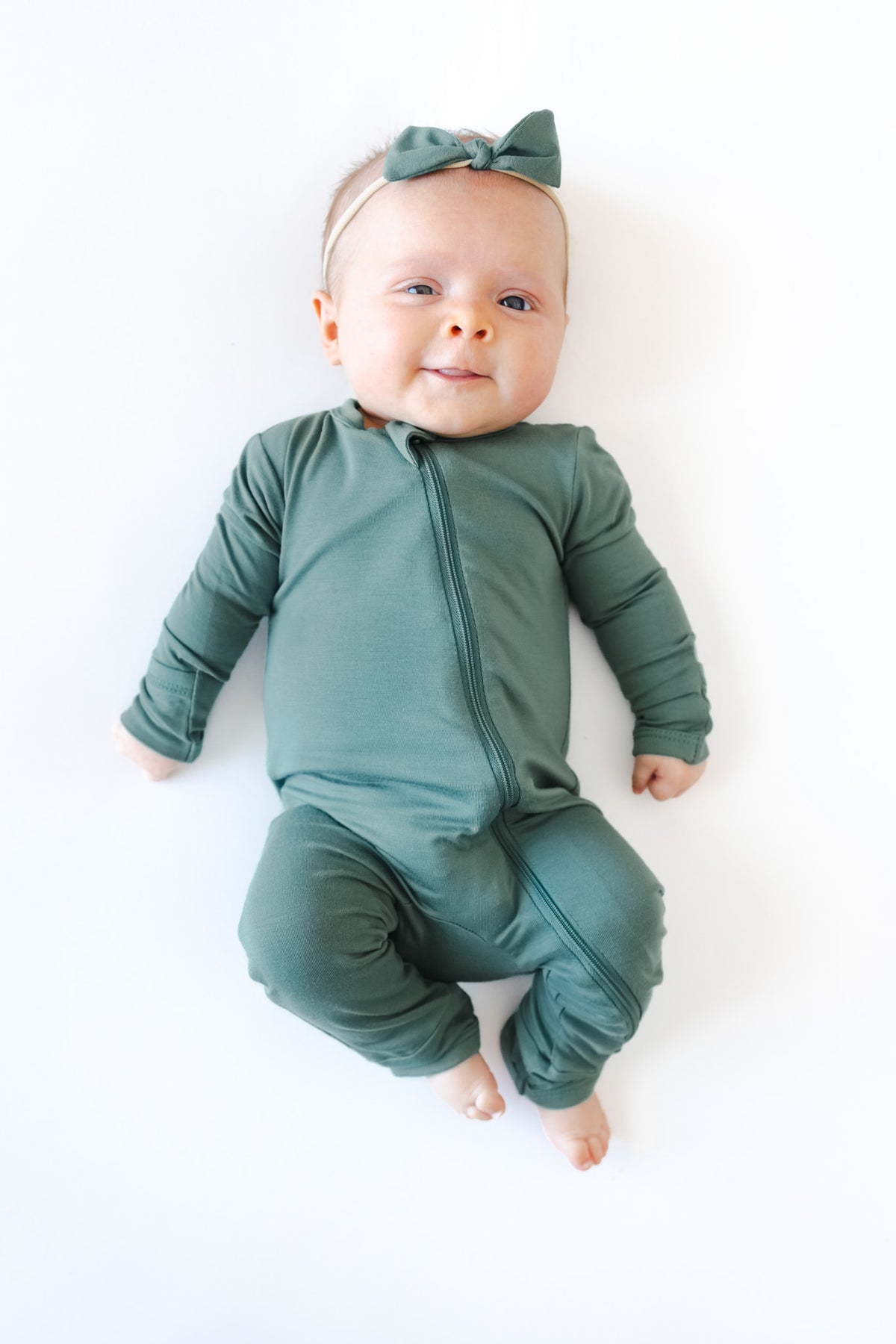 A baby lying on a white background wearing a sage green long-sleeve zip-front romper and a matching bow headband.