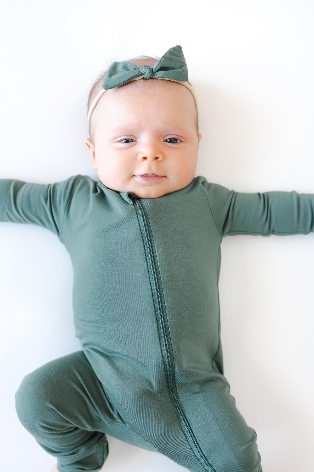 A baby lying on a white background wearing a sage green full-zip footless romper and Tiny Nylon Bow in Pine