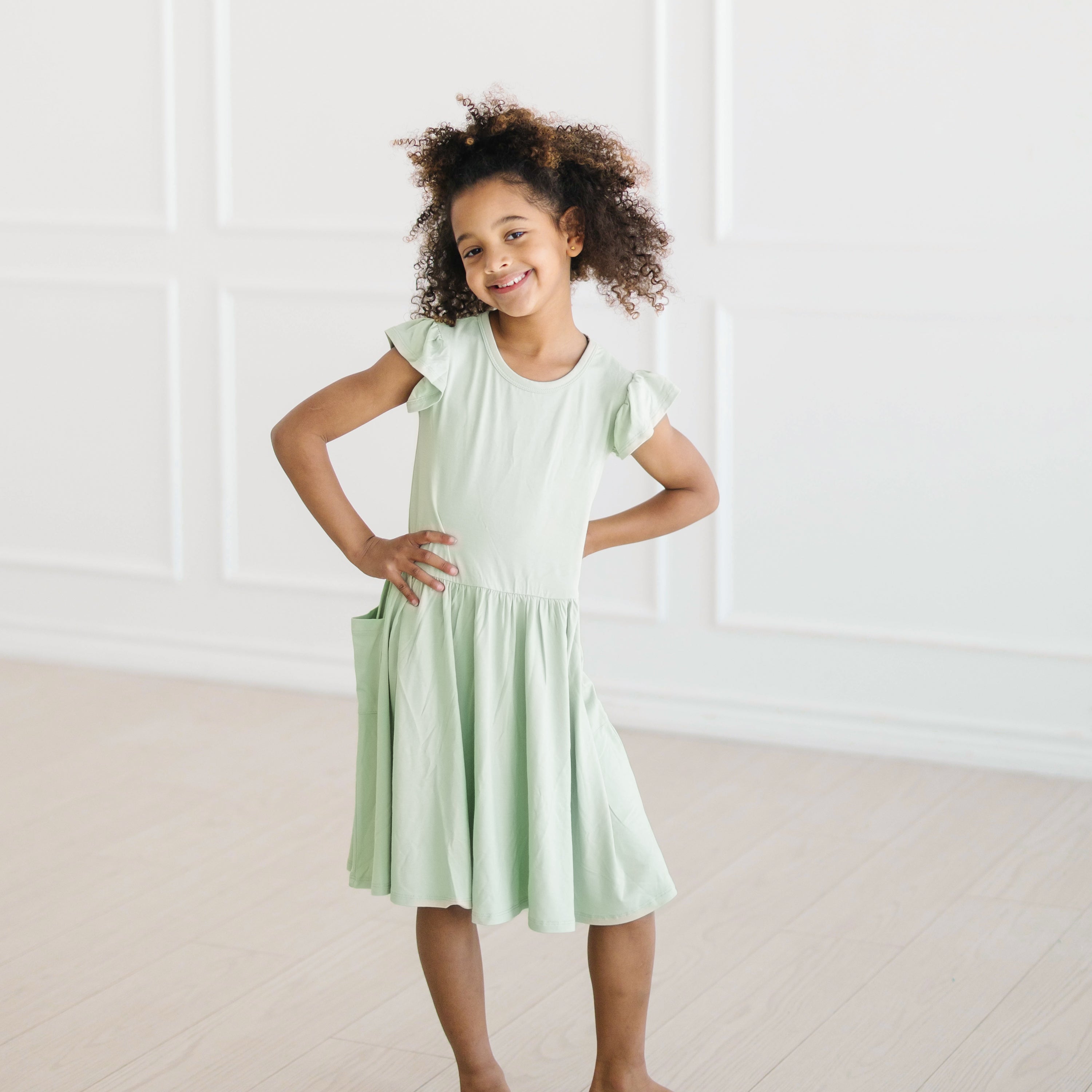 Young girl posing wearing the Pocket Dress in Basil in front of a white wall