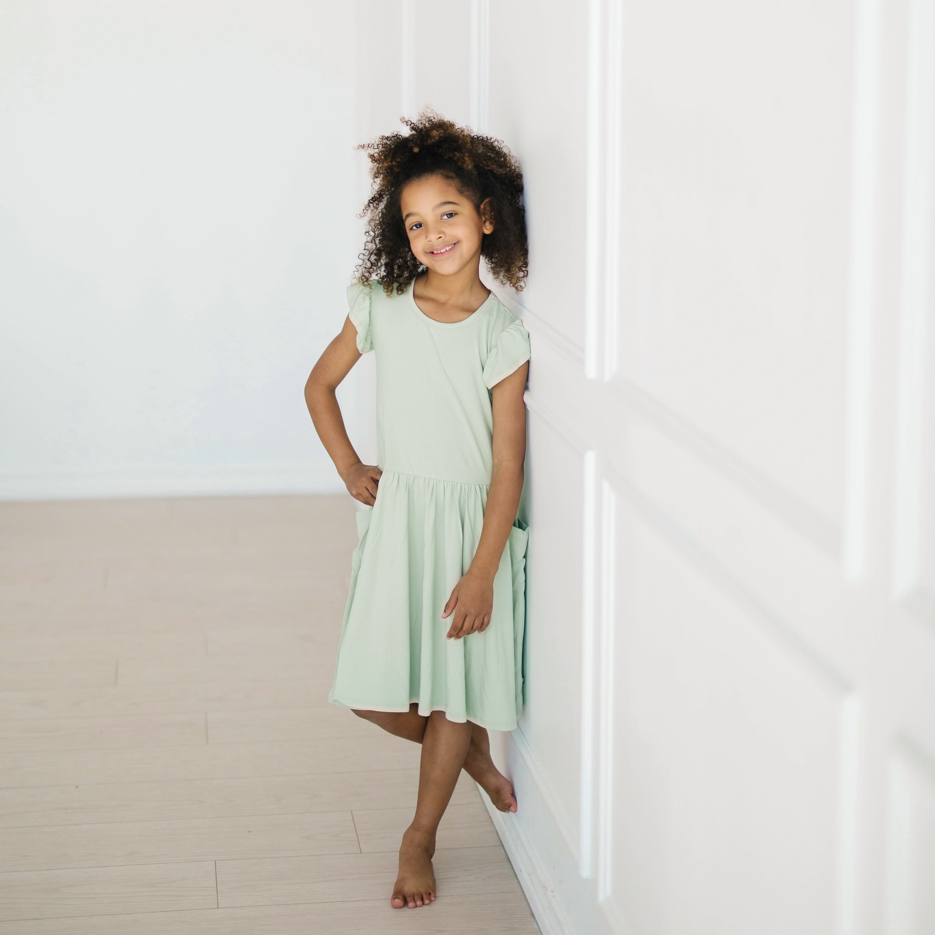 Young girl leaning against a wall wearing the Pocket Dress in Basil