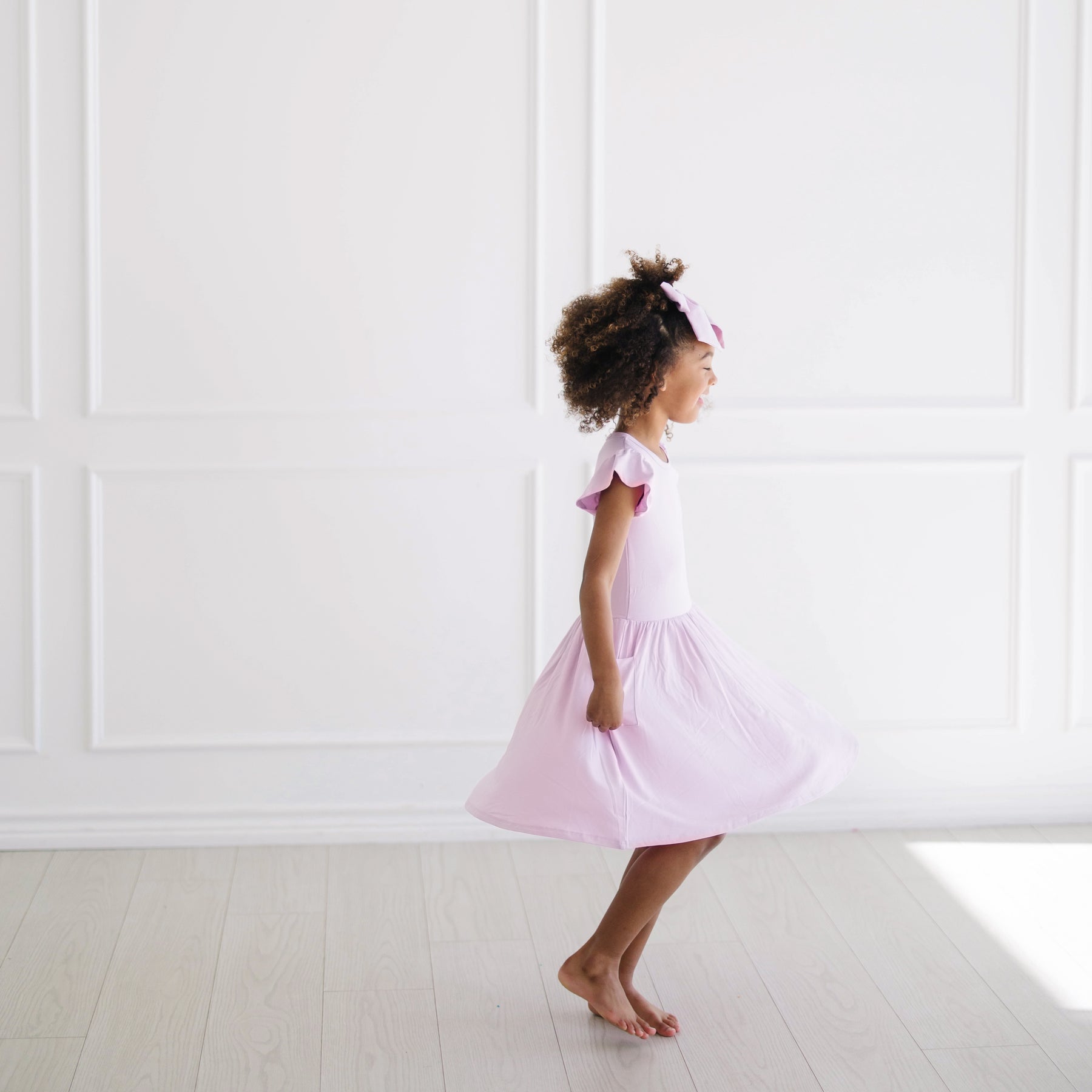 Side view of the Pocket Dress in Thistle on a young girl twirling in front of a white wall
