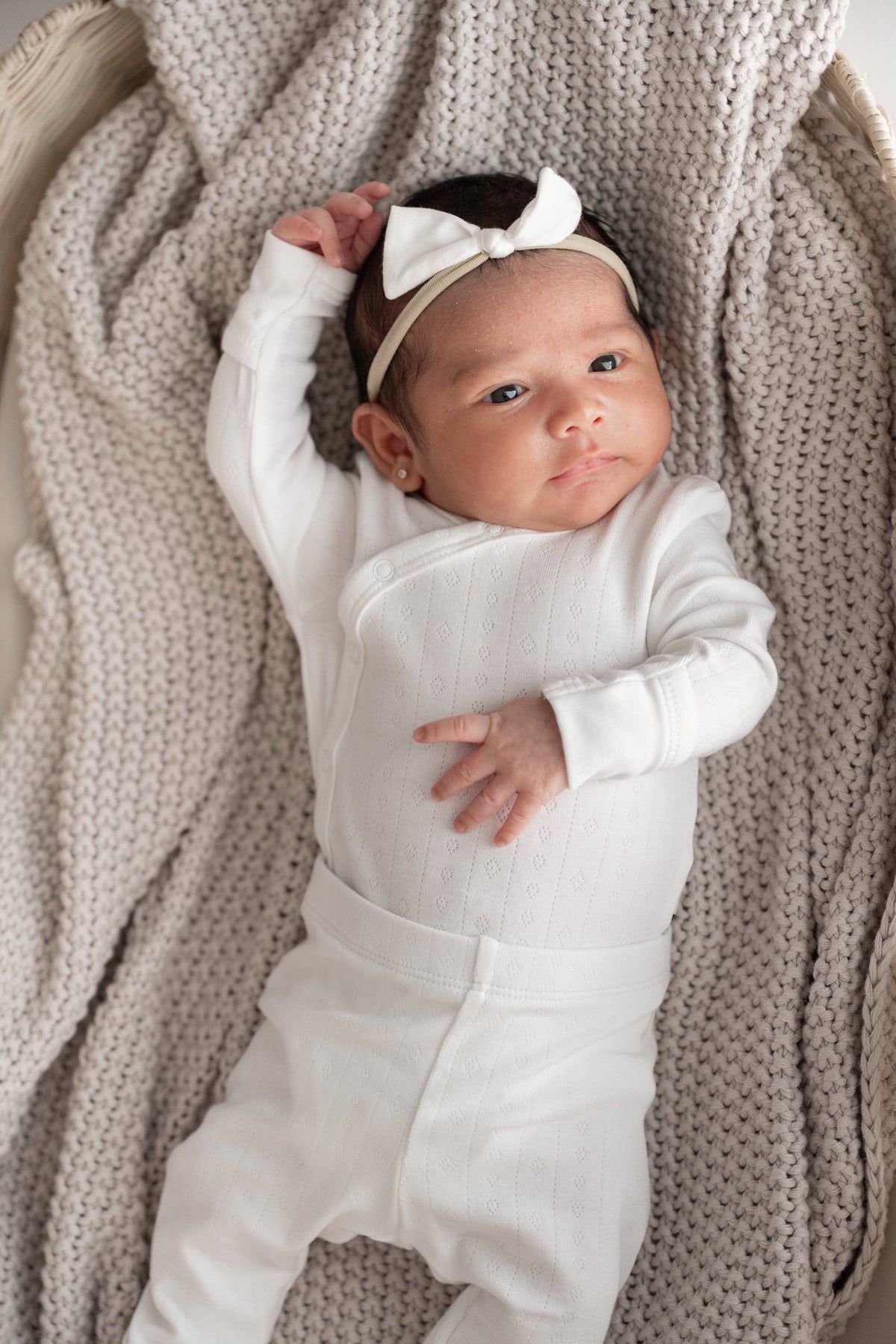 A baby lying on a chunky knit beige blanket, wearing a white long-sleeve bodysuit and a matching white bow headband.