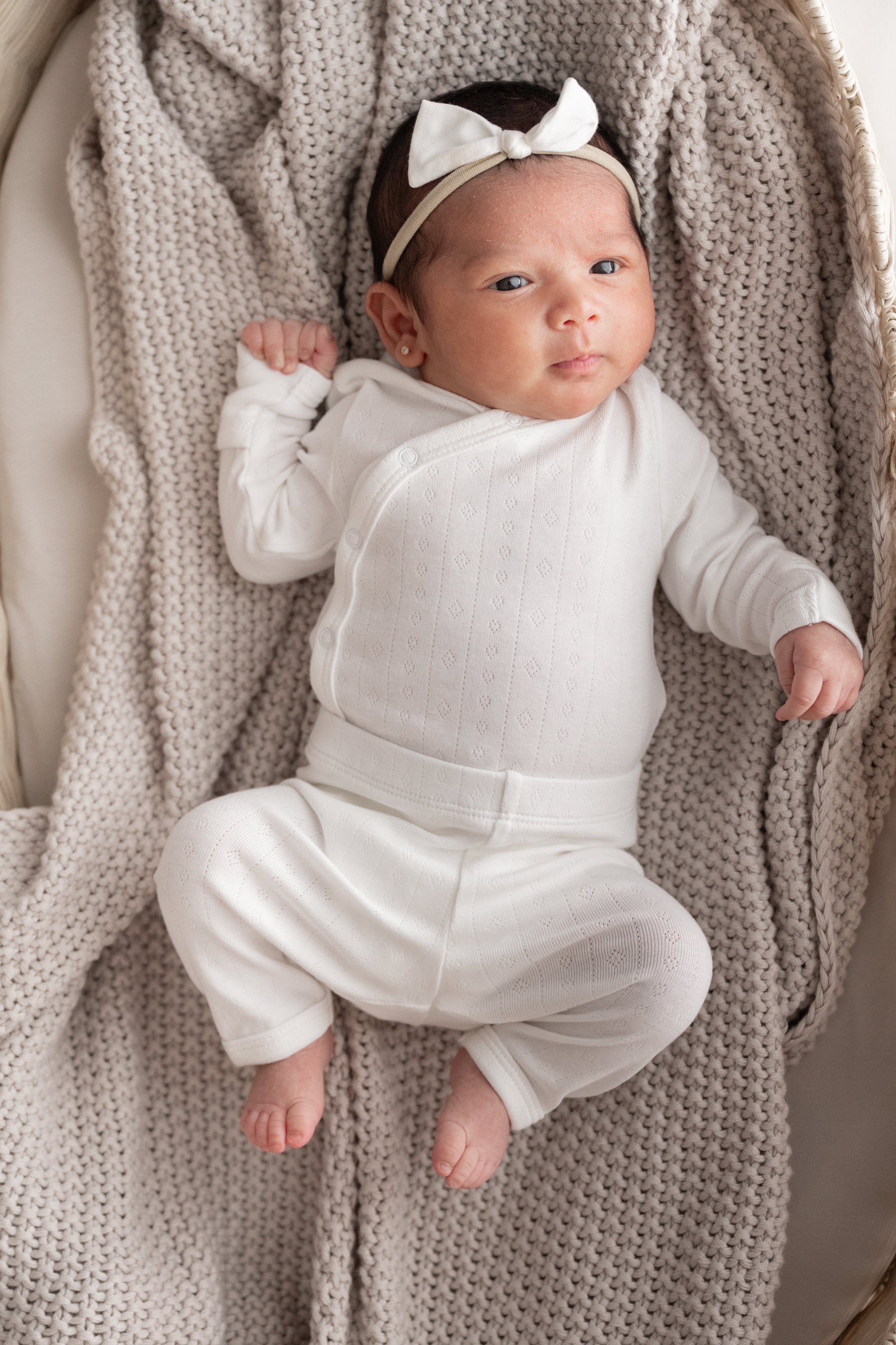 A newborn baby wearing a white long-sleeve wrap bodysuit and matching white bow headband, lying on a textured beige knit blanket.
