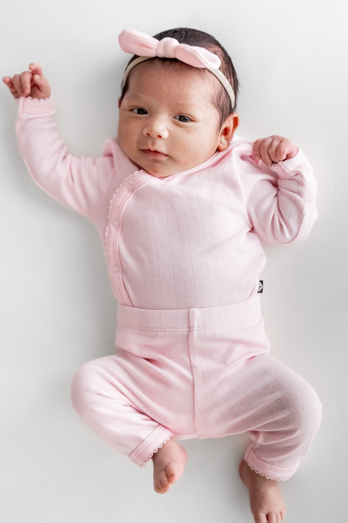 A baby lying on their back wearing a light pink long-sleeve kimono bodysuit and a matching pink bow headband, photographed against a plain light gray background.