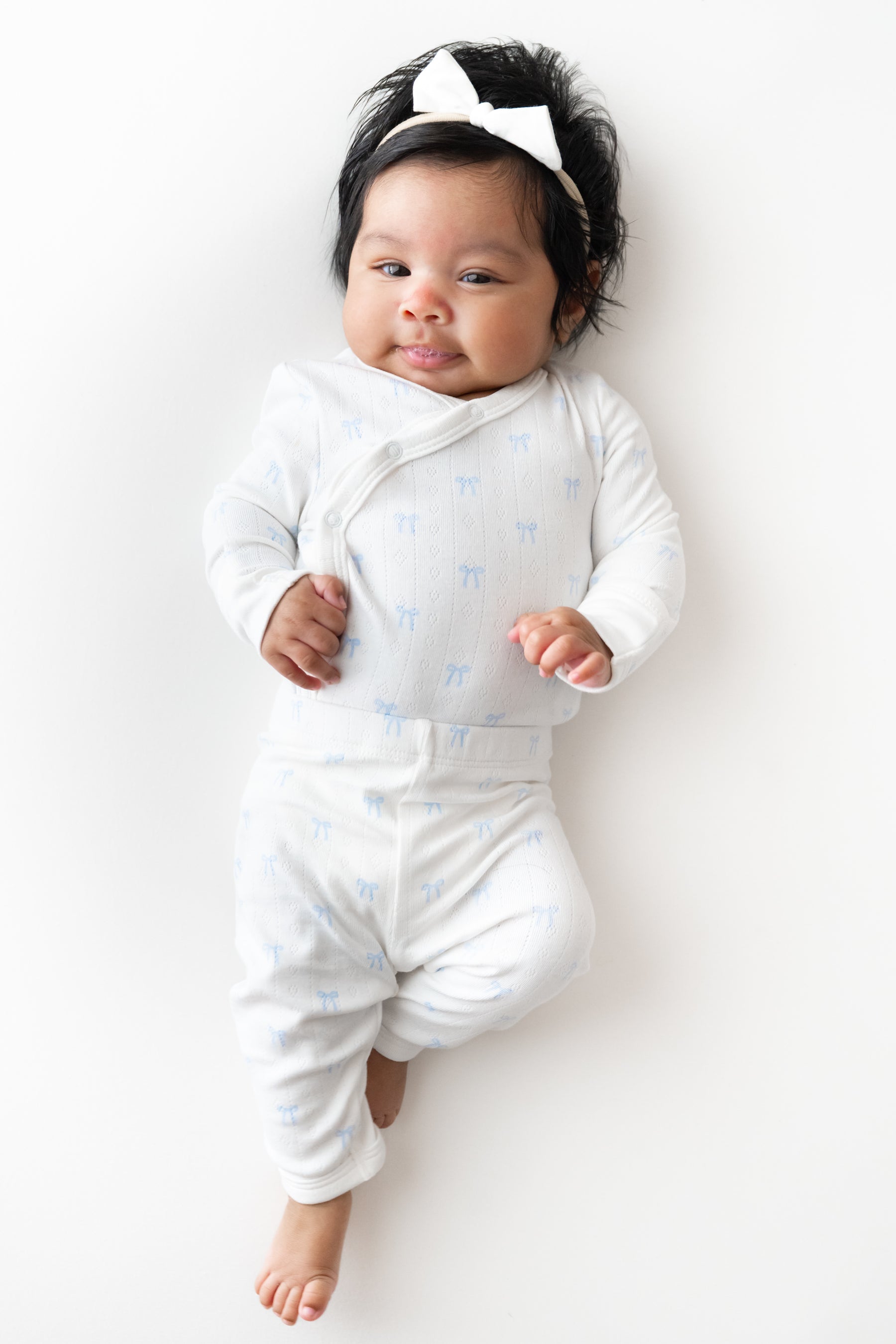 A baby lying on their back wearing a white long-sleeve kimono and pant with a light blue pattern, a white bow headband, and smiling.