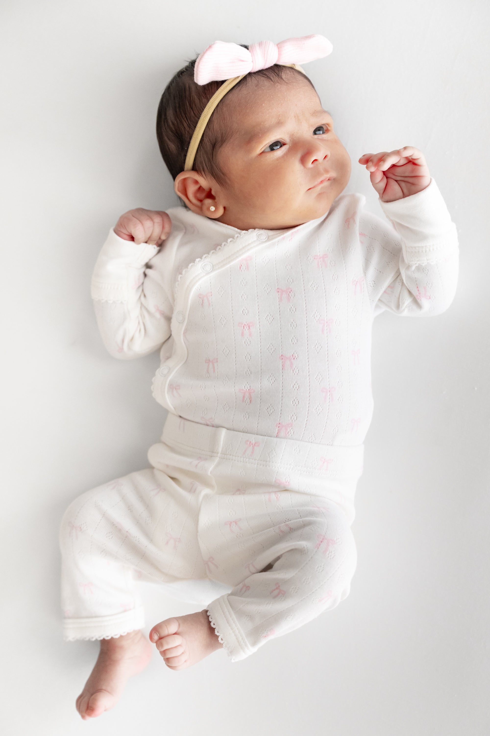 A baby lying on a light gray background wearing a white long-sleeve kimono and pant with tiny pink bow prints, matching white pants, and a pink bow headband.