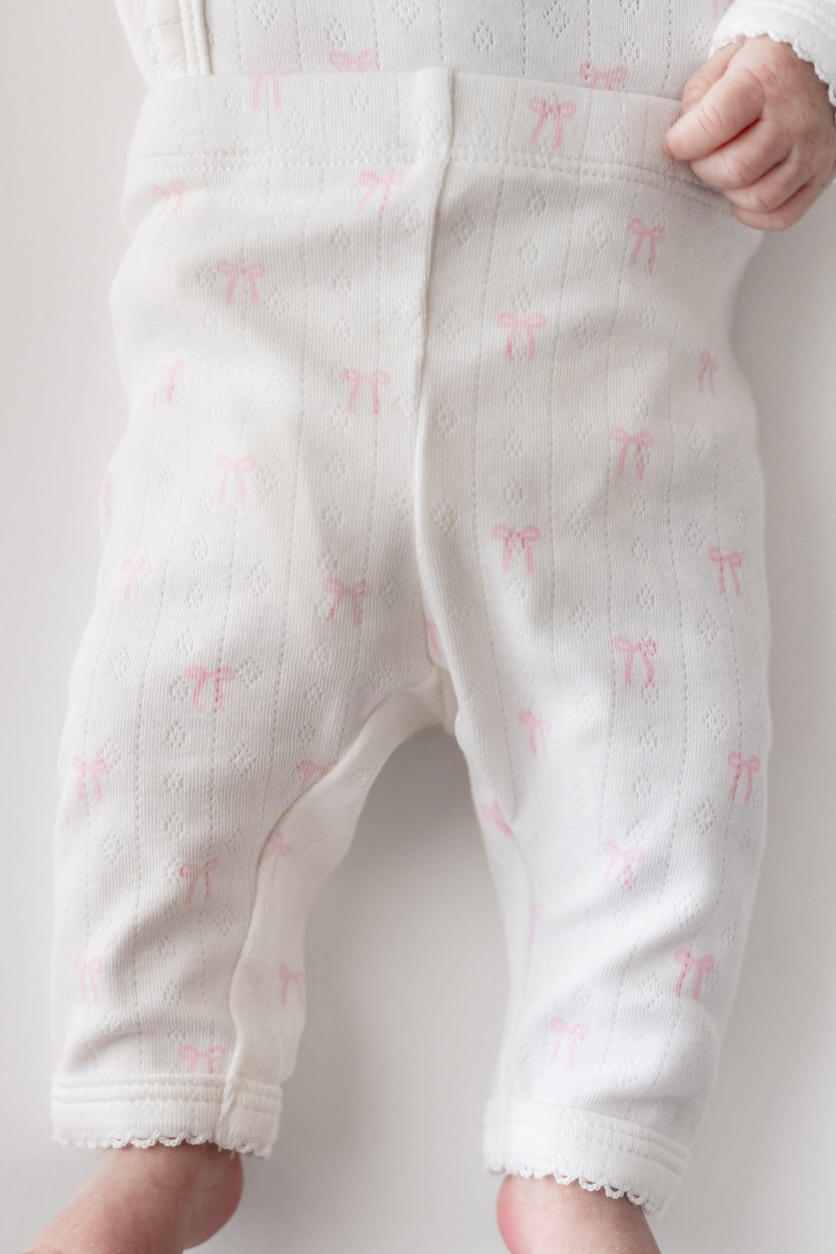 White baby pants with a delicate pink bow pattern, shown on a baby with a hand visible at the waistband.