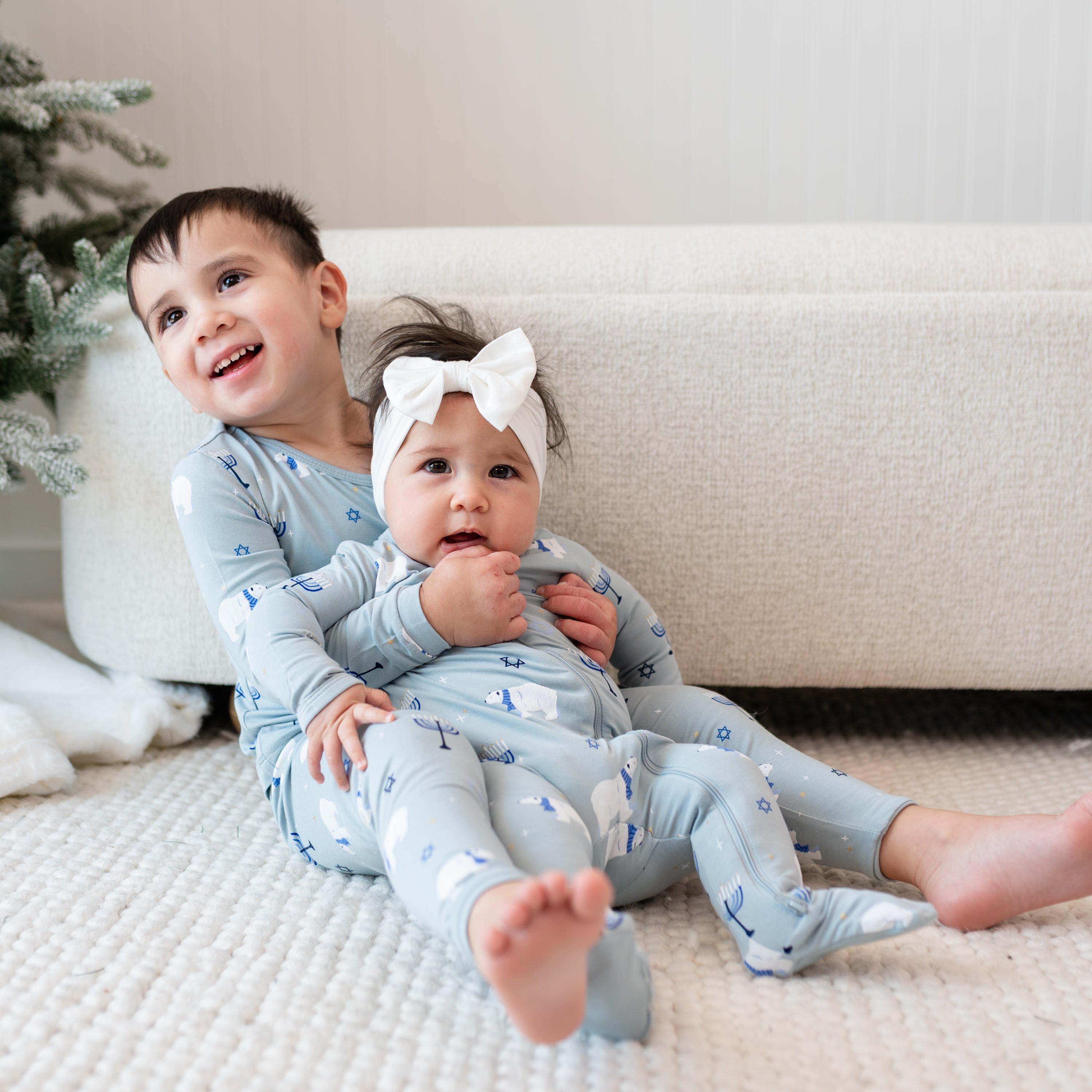 Young boy sitting wearing the Long Sleeve Pajamas in Polar Lights holding his younger sister who is matching him in a zippered footie and Cloud bow headband