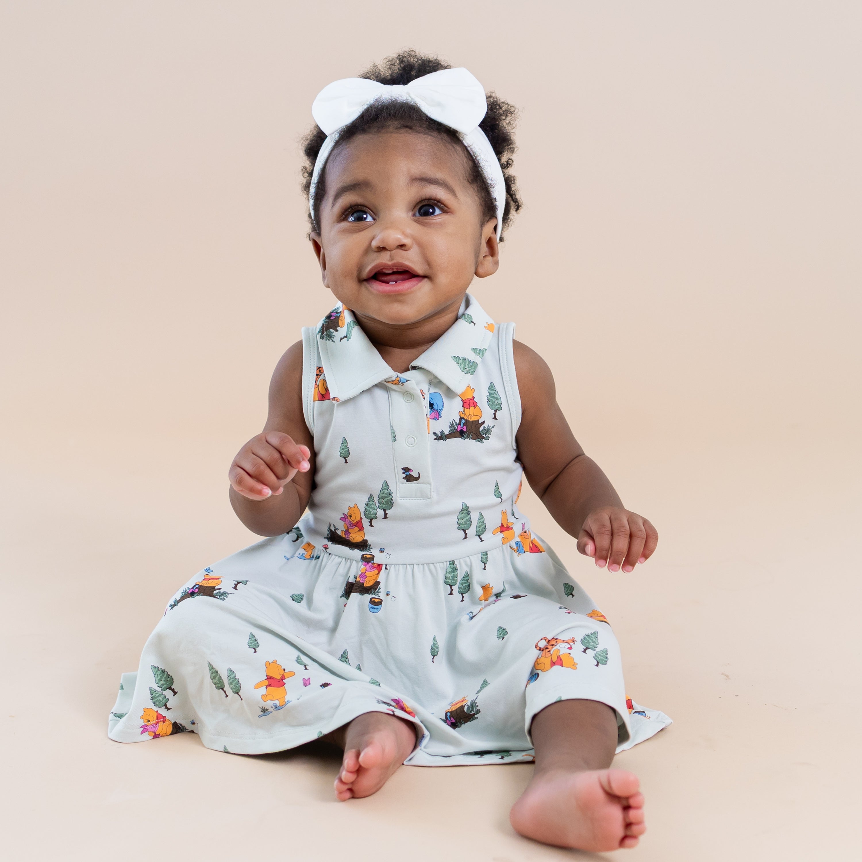 Toddler sitting on the floor wearing the Polo Bodysuit Dress in Winnie the Pooh Hundred Acre Wood paired with a white bow