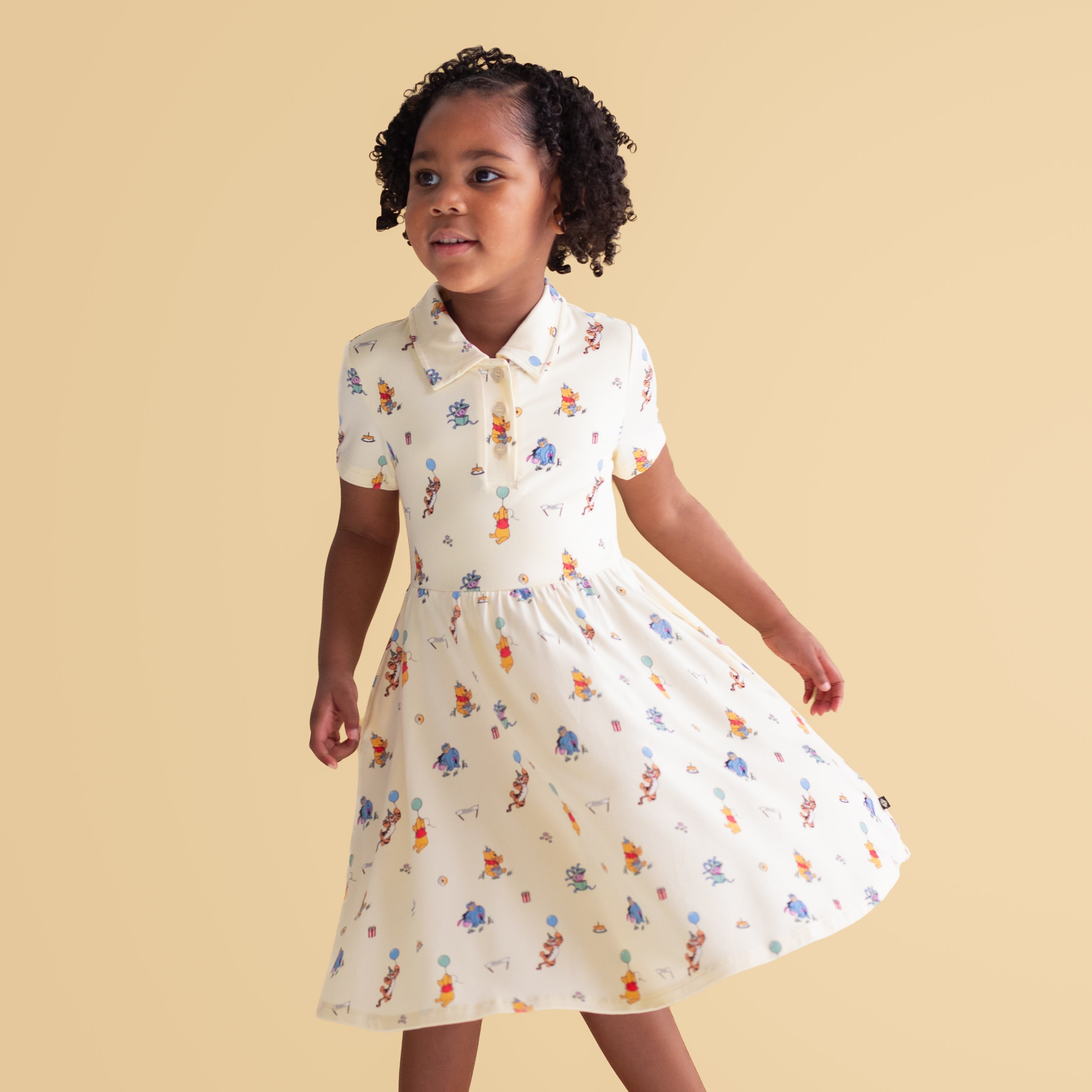Young girl walking wearing the Polo Dress in Winnie the Pooh Celebration in front of a mustard yellow background