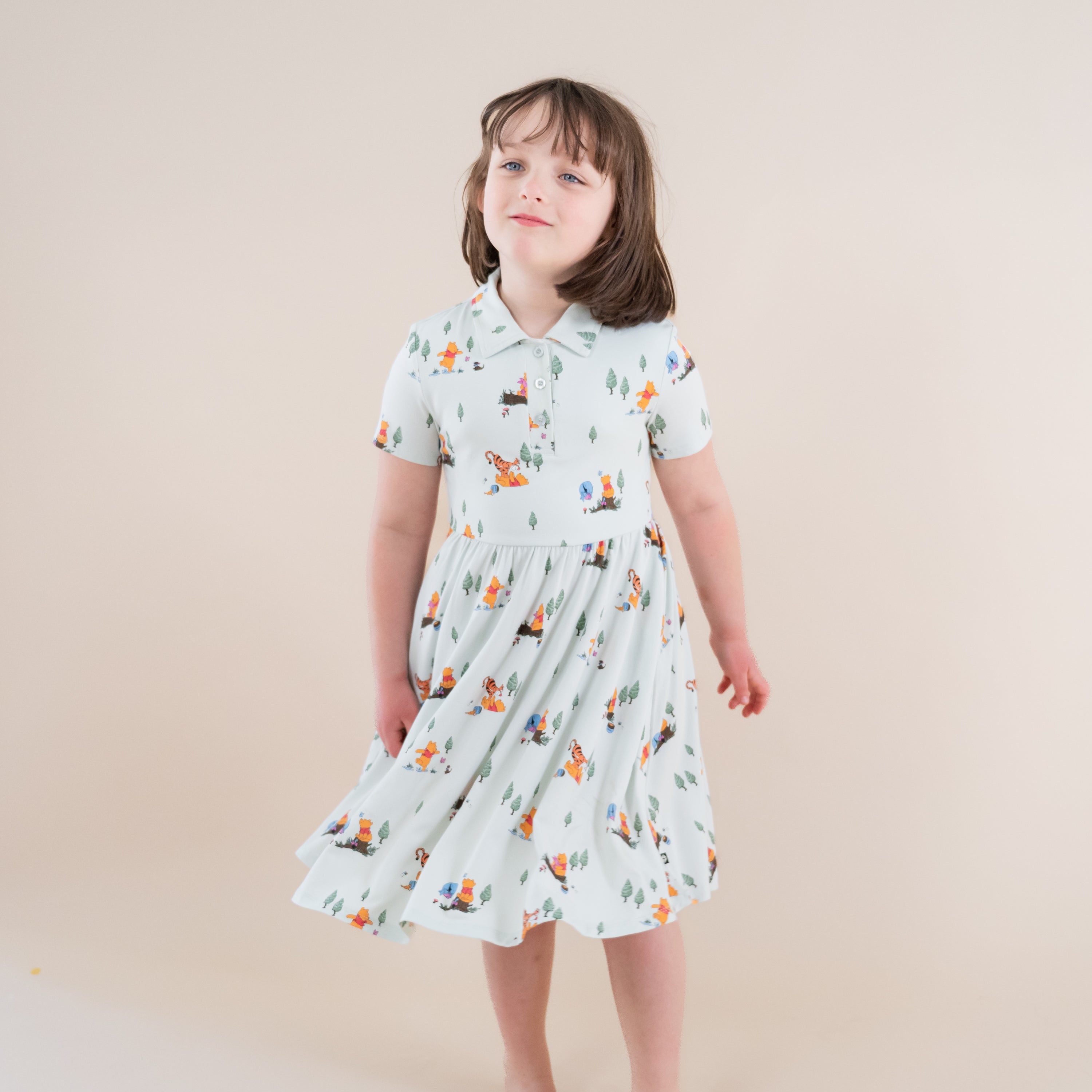 Young girl standing in front of a light neutral beige wall wearing the Polo Dress in Winnie the Pooh Hundred Acre Wood