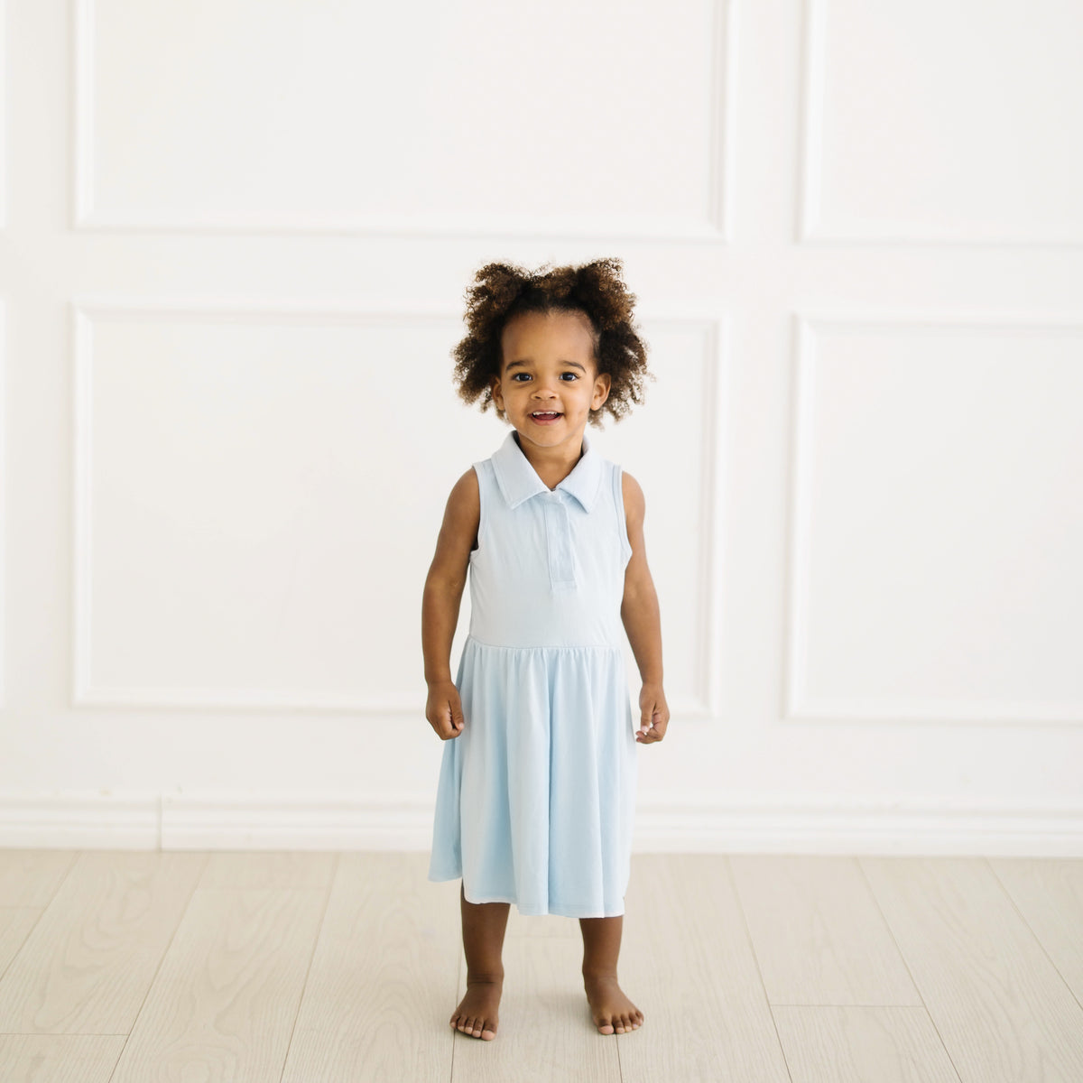 Young toddler standing in front of a white wall wearing the Polo Bodysuit Dress in Breeze