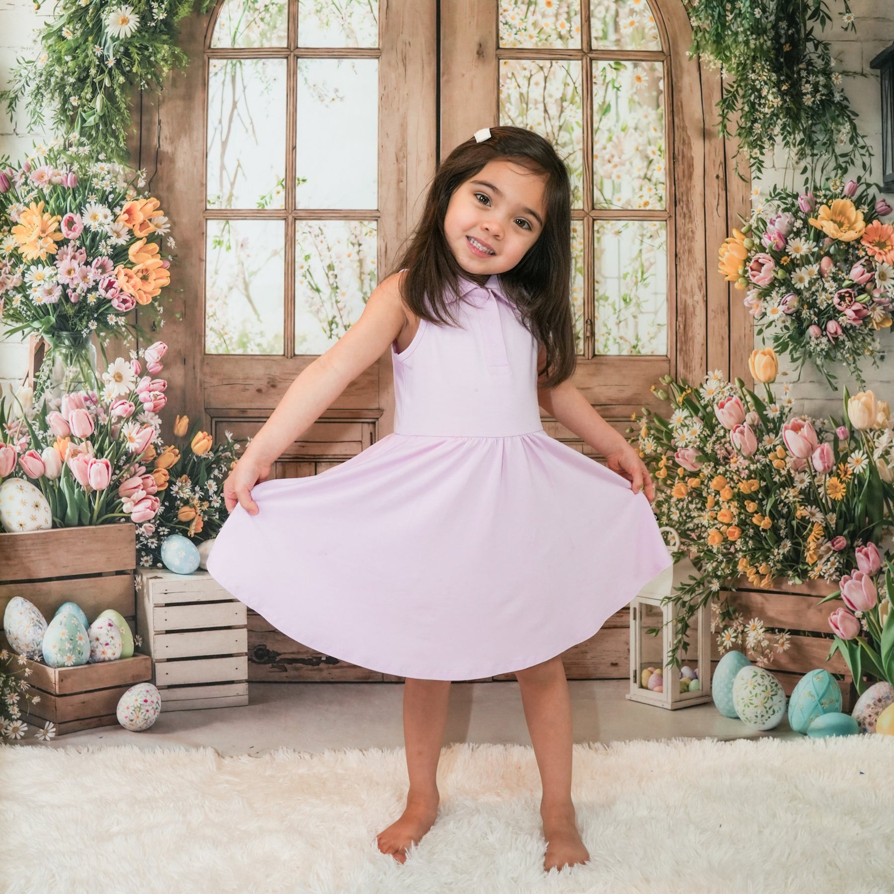 Smiling girl standing in front of a wooden window with flowers holding the hem of the skirt of the Polo Bodysuit Dress in Thistle 