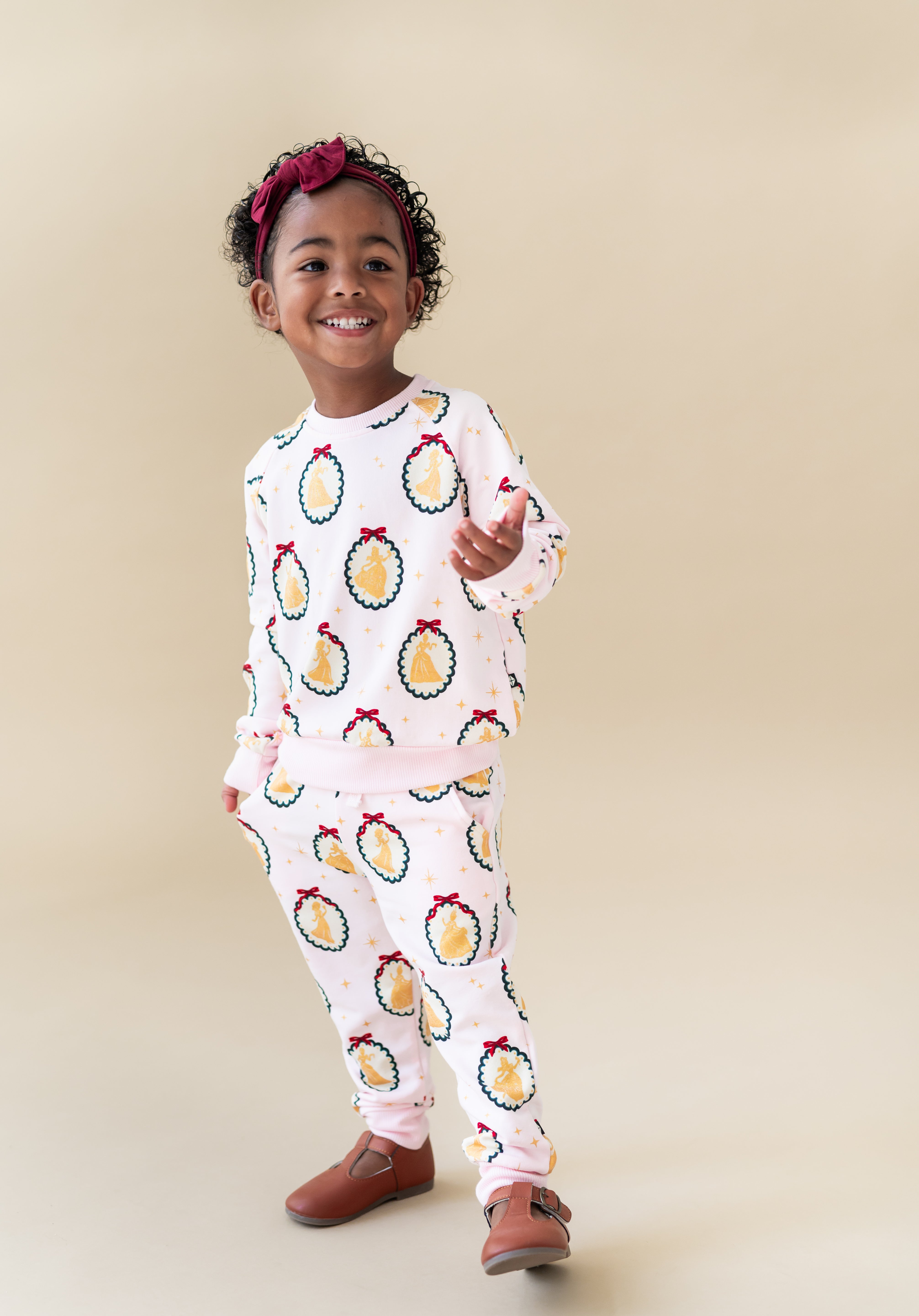 Child posing in Long Sleeve Jogger Set in Princess Holiday Portraits print and knotted bow headband on a beige background