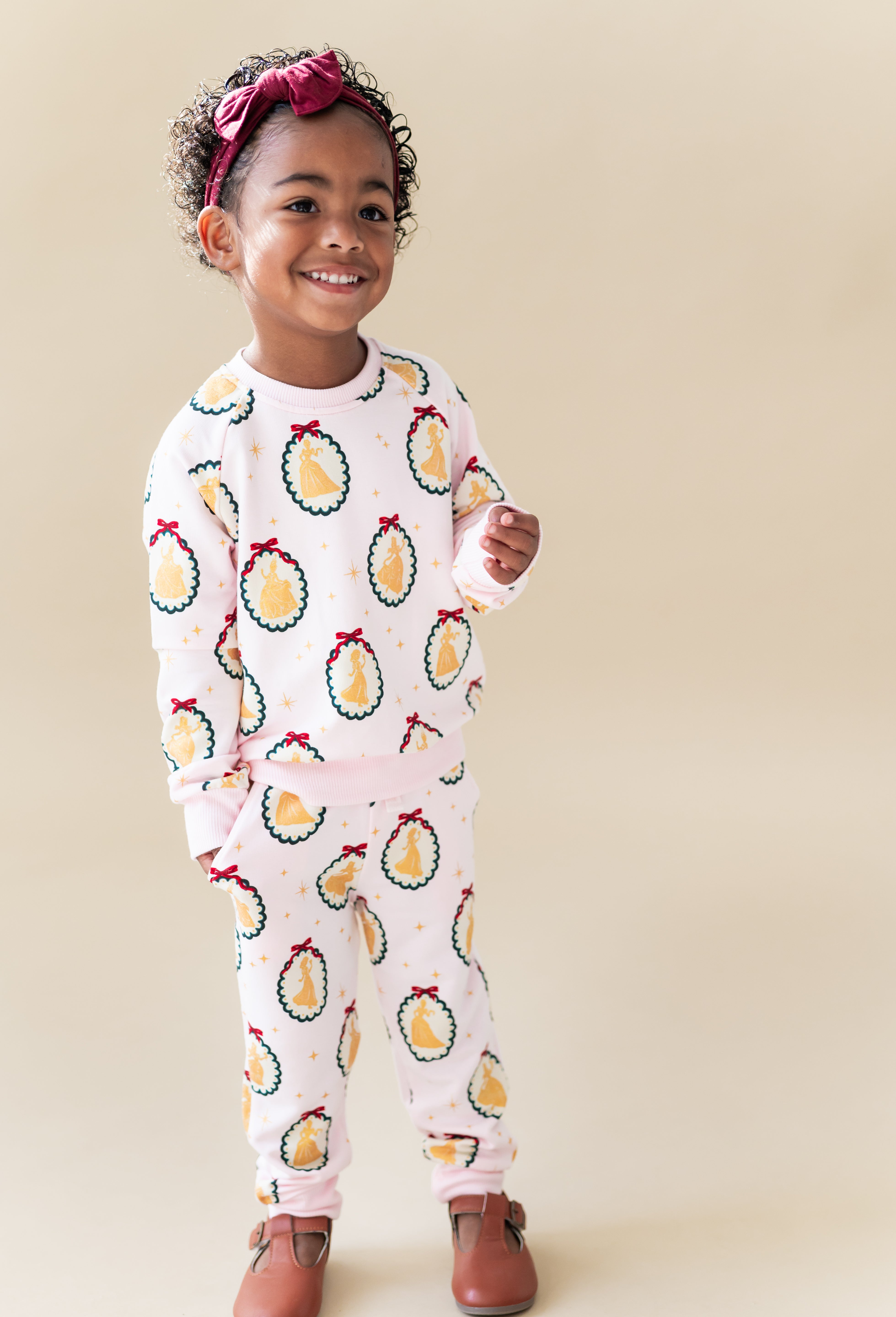 Child smiling in Long Sleeve Jogger Set in Princess Holiday Portraits print and knotted bow headband on a beige background