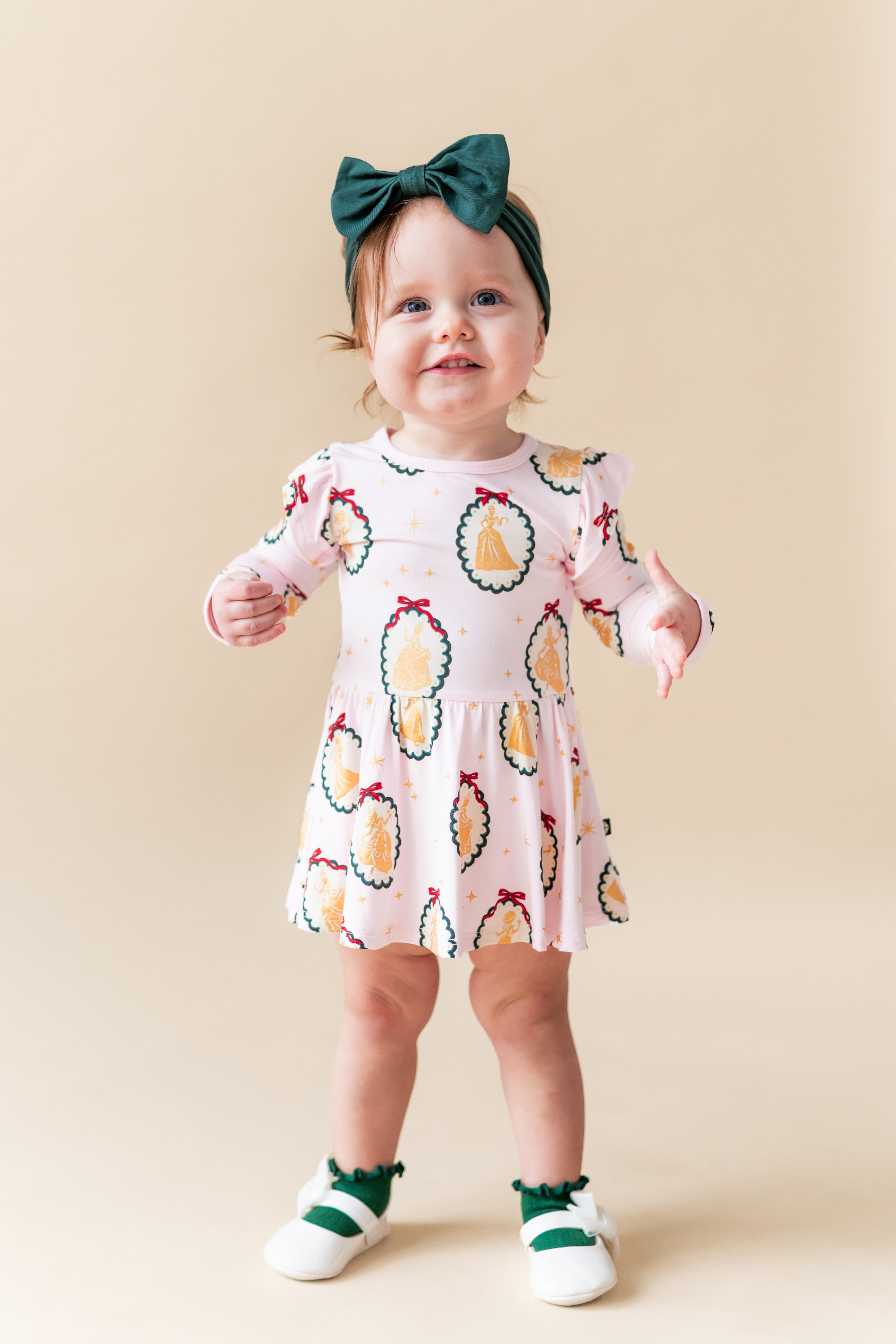 Girl wearing Long Sleeve Twirl Bodysuit Dress in Princess Holiday Portraits print with green knotted bow on a beige background