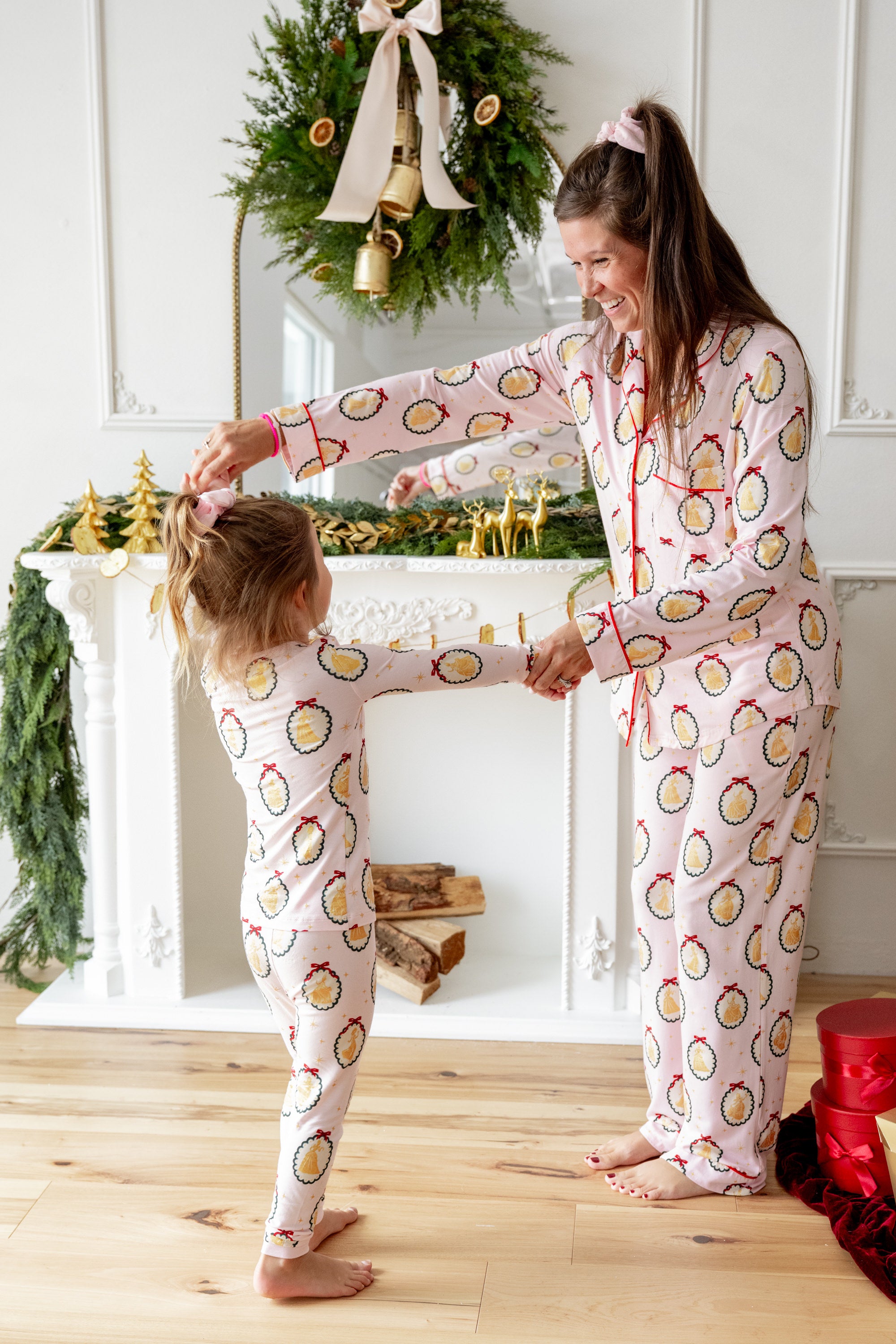 Mommy and Toddler wearing matching print Long-Sleeved Pajama Set in Princess Holiday Portraits print standing in front of fire place decorated with Christmas thyme dancing along