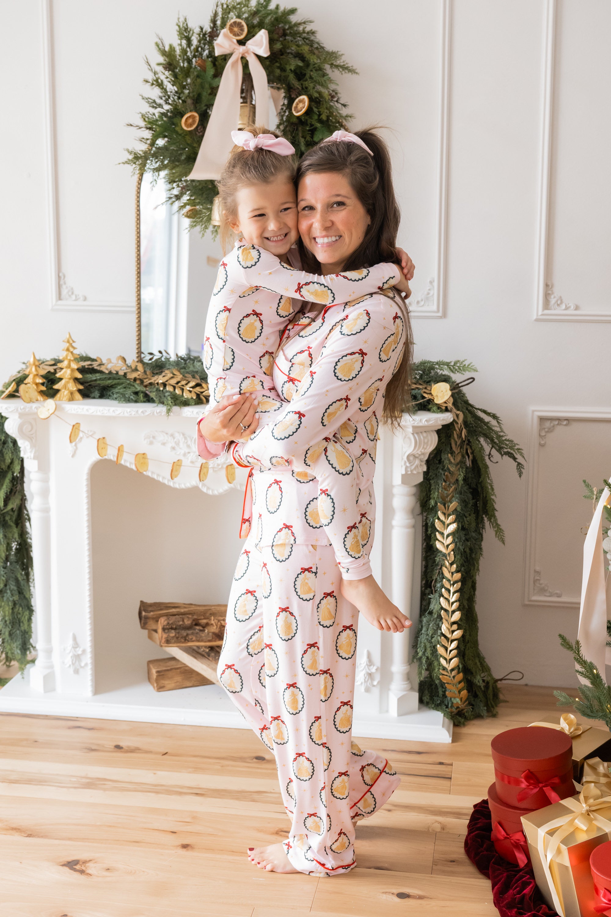 Mommy and Toddler wearing matching print Long-Sleeved Pajama Set in Princess Holiday Portraits print standing in front of fire place decorated with Christmas thyme