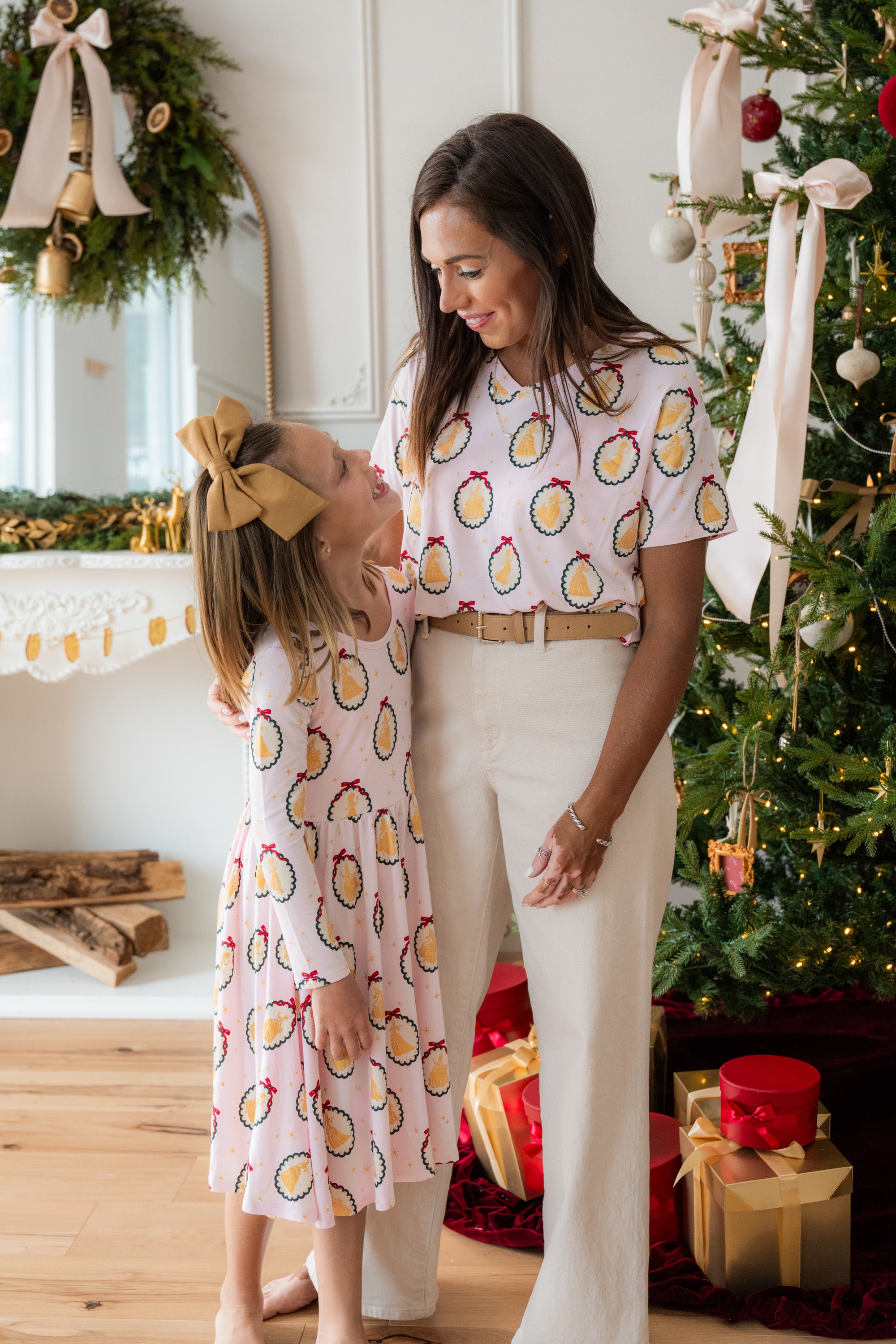 Woman and girl wearing matching outfits wearing Women's Oversized V-Neck Tee in Princess Holiday Portraits print over white trousers and long sleeve skater dress in princes holiday portraits in front of fire place and presents