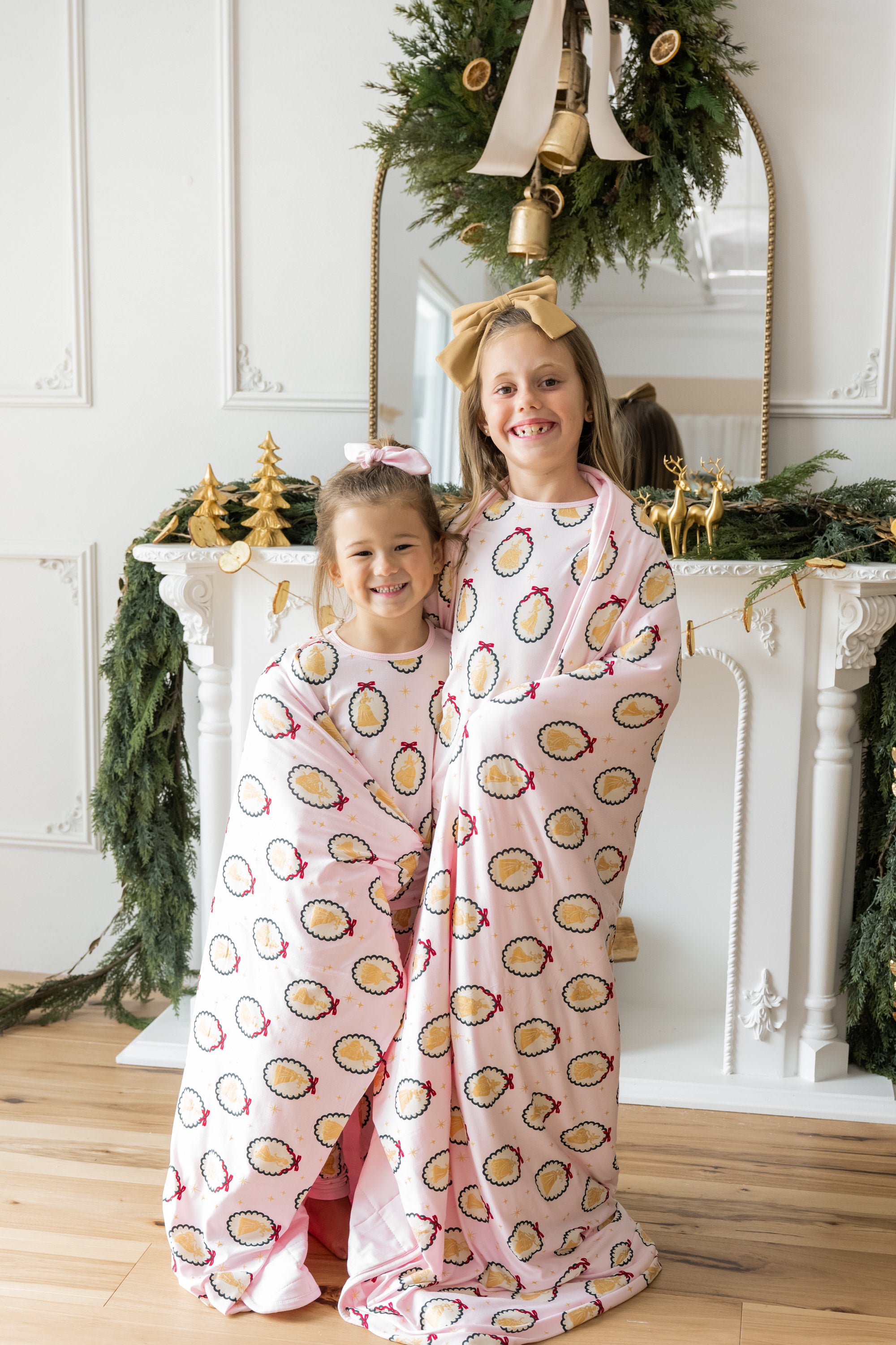 Two girls wrapped in Youth Blanket in Princess Holiday Portraits 2.5 print smiling together in front of fire place decorated in Christmas thyme