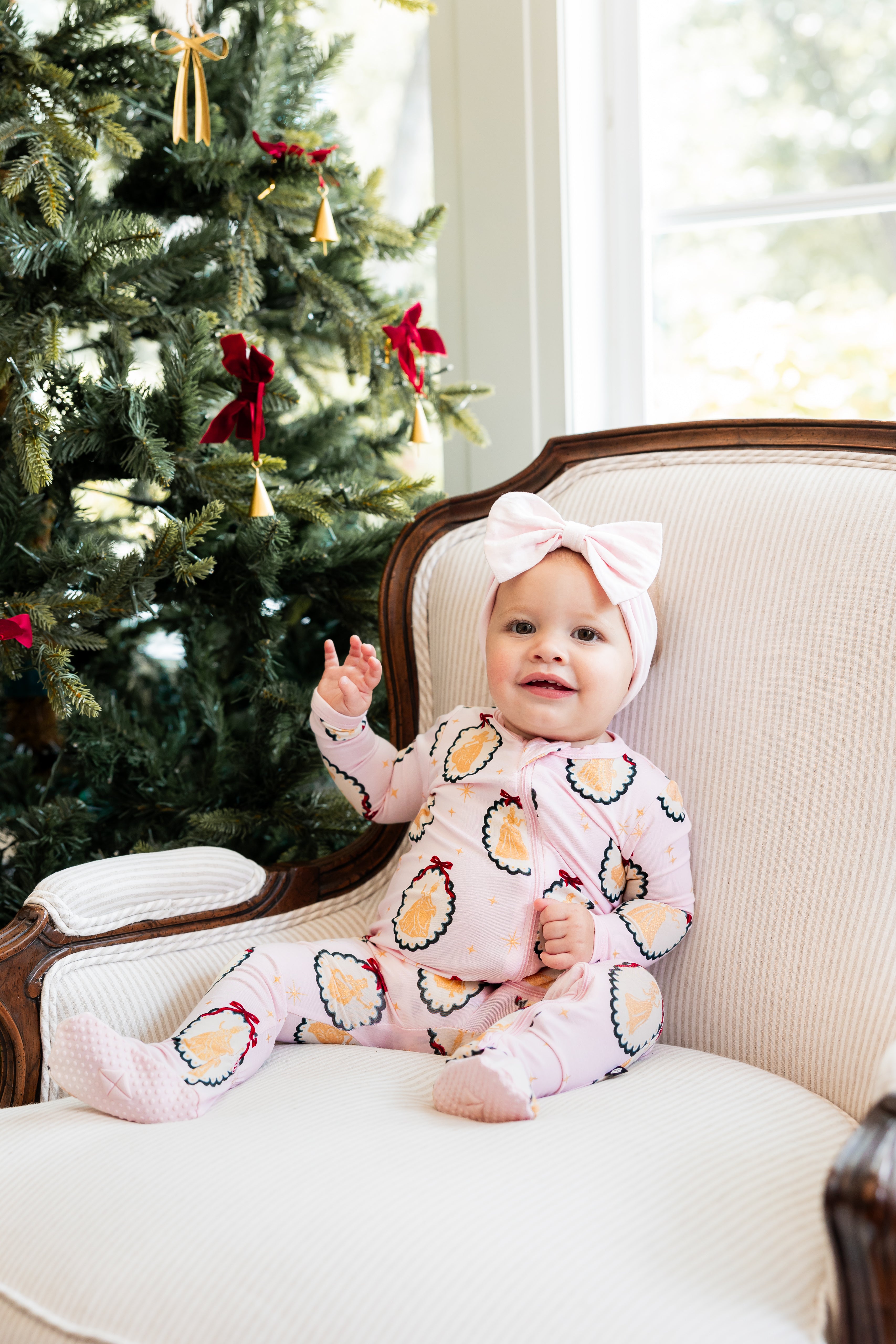 Baby sitting on sofa wearing Zippered Footie in Princess Holiday Portraits print with knotted bow headband beside a Christmas tree