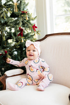Baby wearing Zippered Footie in Princess Holiday Portraits print sitting on sofa beside a Christmas tree